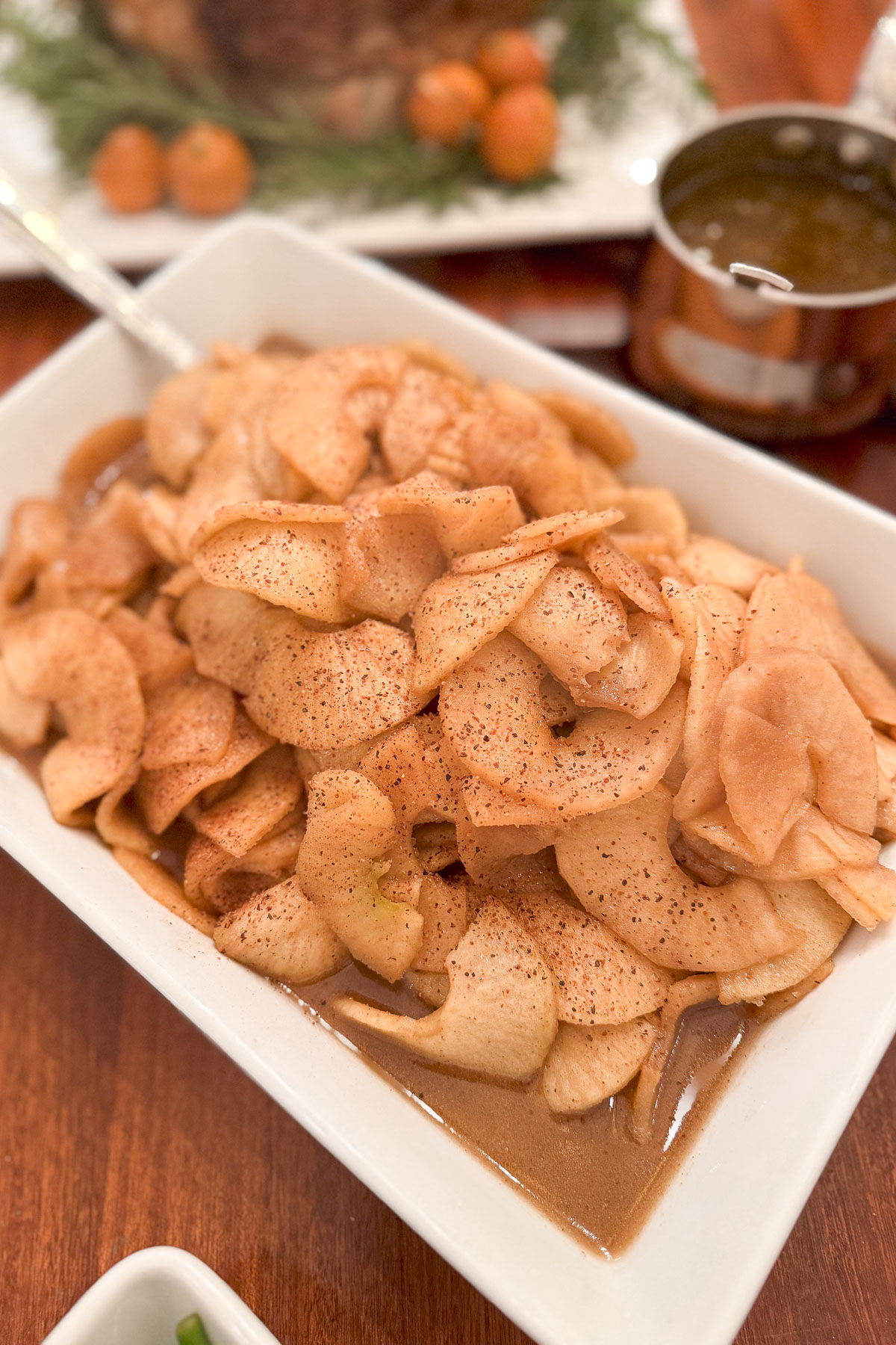 A rectangular white dish filled with sliced cooked apples, seasoned with cinnamon and sitting in a light brown sauce. A serving spoon is partially visible on the side. There's a blurred background with more food and a small pot.