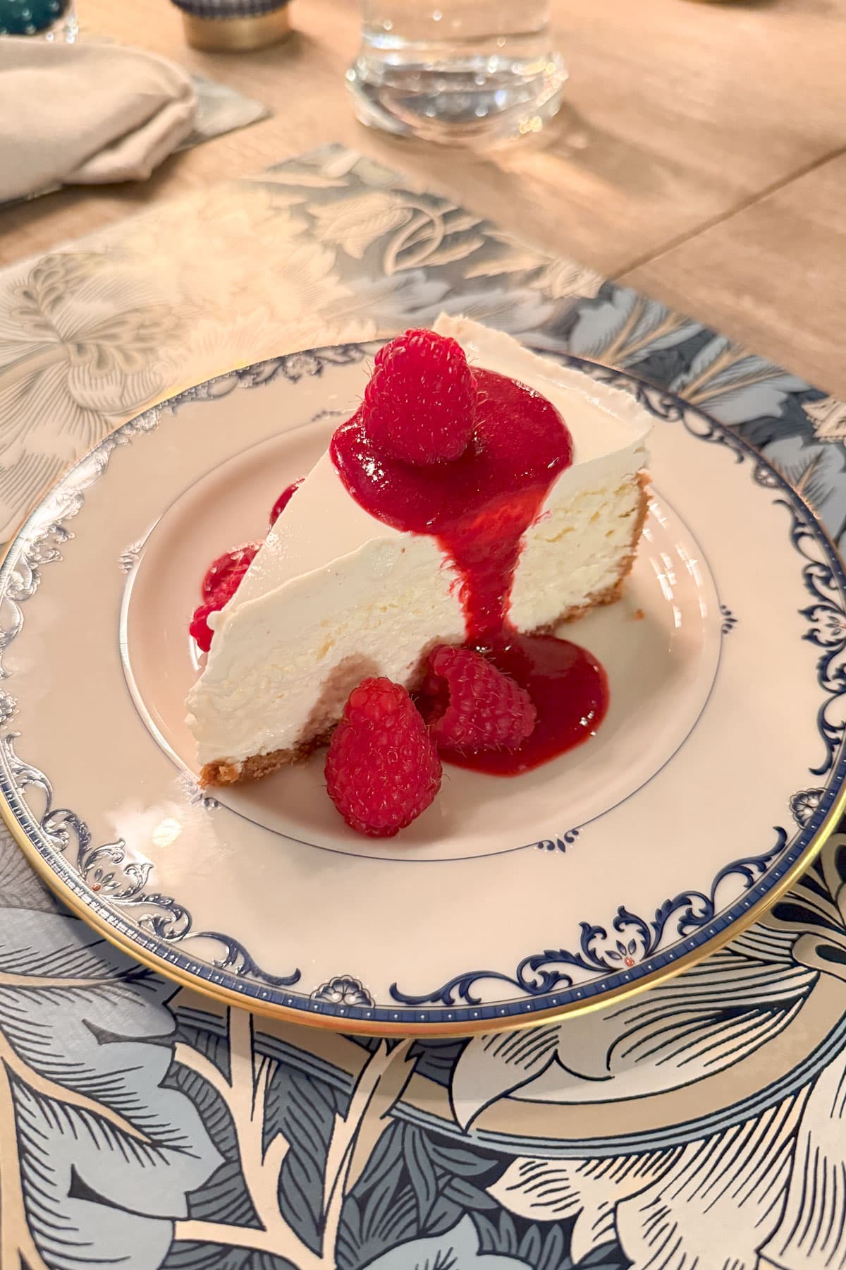 A slice of cheesecake topped with bright red raspberry sauce and fresh raspberries graces a decorative blue and white plate, perfect for Sunday Sharing. Resting on a wooden table adorned with a patterned tablecloth, this delightful treat invites moments of indulgence.