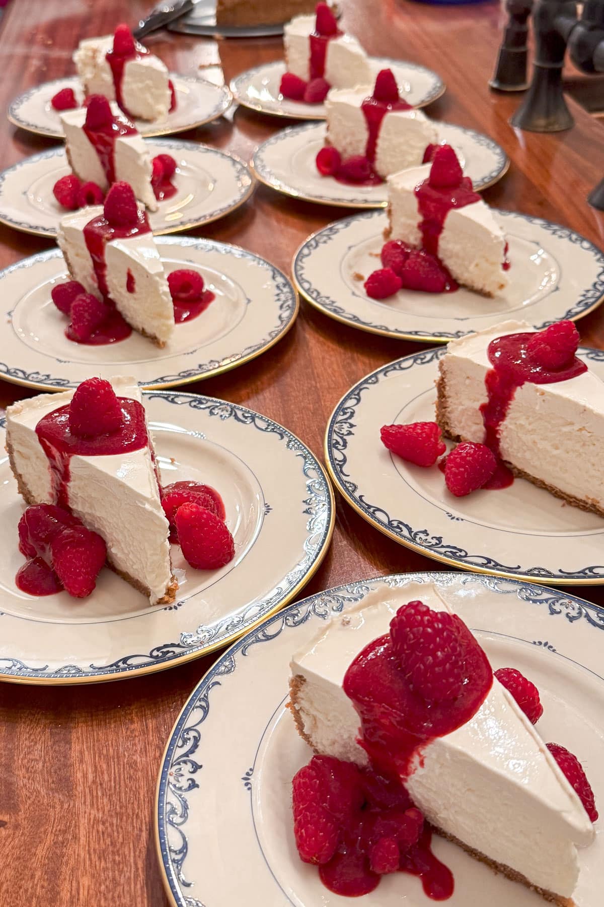 Slices of cheesecake topped with raspberry sauce and fresh raspberries are served on white plates with a blue design, arranged in rows on a wooden table.