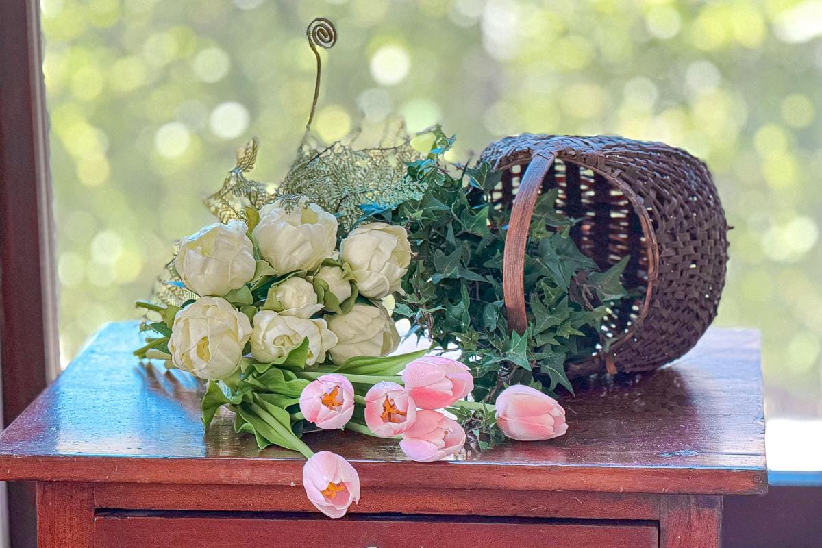 A bouquet of white flowers and green leaves sits on a wooden table. Nearby, a wicker basket is tipped over, spilling pink tulips. The background shows blurred greenery through a window.