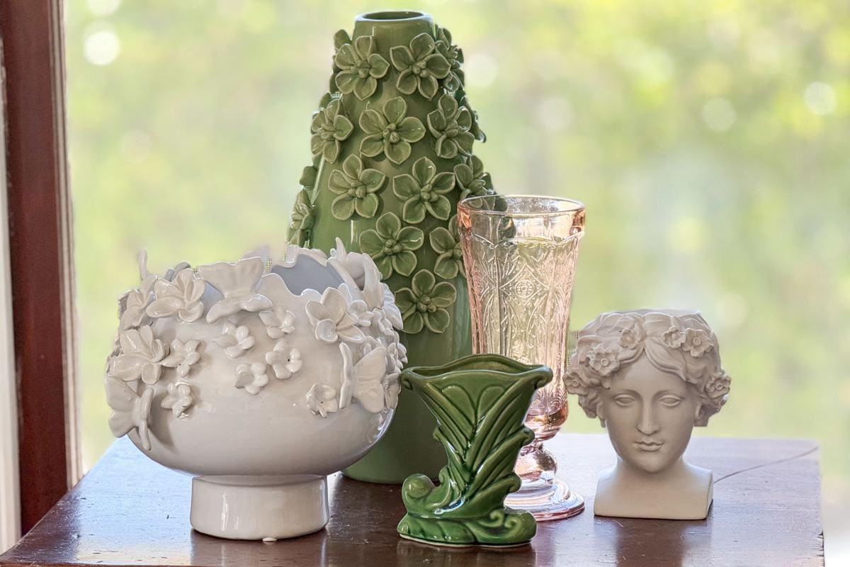 A group of decorative vases and sculptures on a table. A white bowl with floral design, a tall green vase with flowers, an ornate pink glass, a small green leaf-shaped vase, and a white bust with floral headpiece are arranged together.