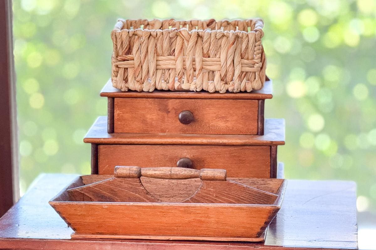 A wooden table holding a two-tier wooden chest with small drawers. On top of the chest is a woven straw basket. In front of the chest is a wooden tray with handles. The background is softly blurred with green and yellow tones.