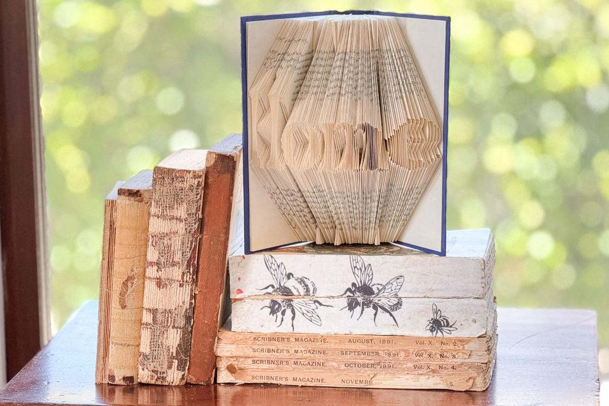 A stack of vintage books is topped with an open book, its pages artistically folded to spell "home." The image is brightly lit with a blurred green background, suggesting a window view.