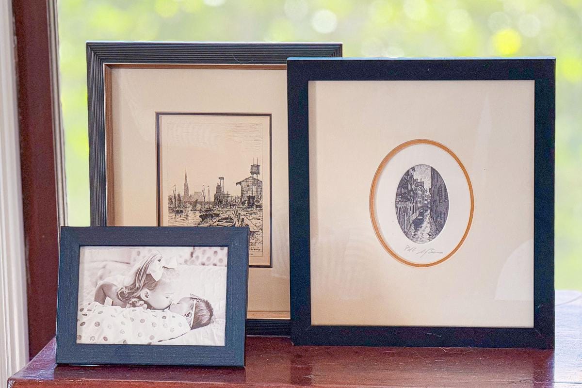 Three framed pictures on a wooden surface. The first is a black and white photo of a baby and child. The second is a cityscape sketch with buildings and water towers. The third is an oval-shaped vintage illustration in a black frame.