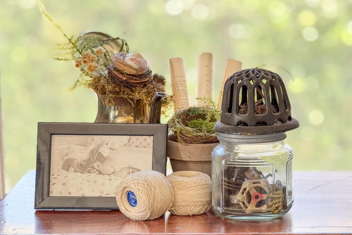 A rustic arrangement on a wooden table featuring a framed photo, a vintage teapot with moss and flowers, a pot with rolled papers and moss, twine balls, gears in a jar, and a decorative metal cover. Blurred green background.