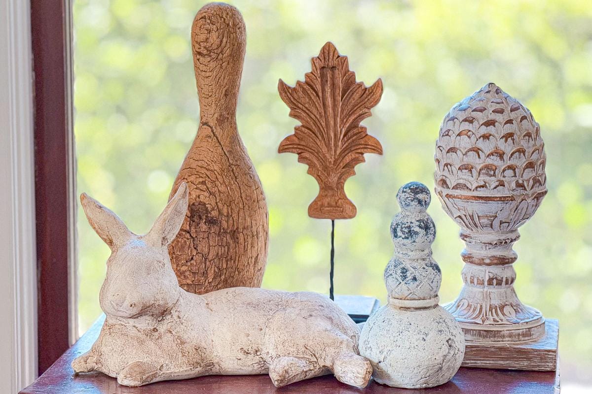 A collection of decorative wooden and stone ornaments on a surface. Includes a rabbit, a bowling pin shape, a leaf design, a carved pinecone, and a snowman shape. The background is a blurred outdoor scene with green and yellow tones.