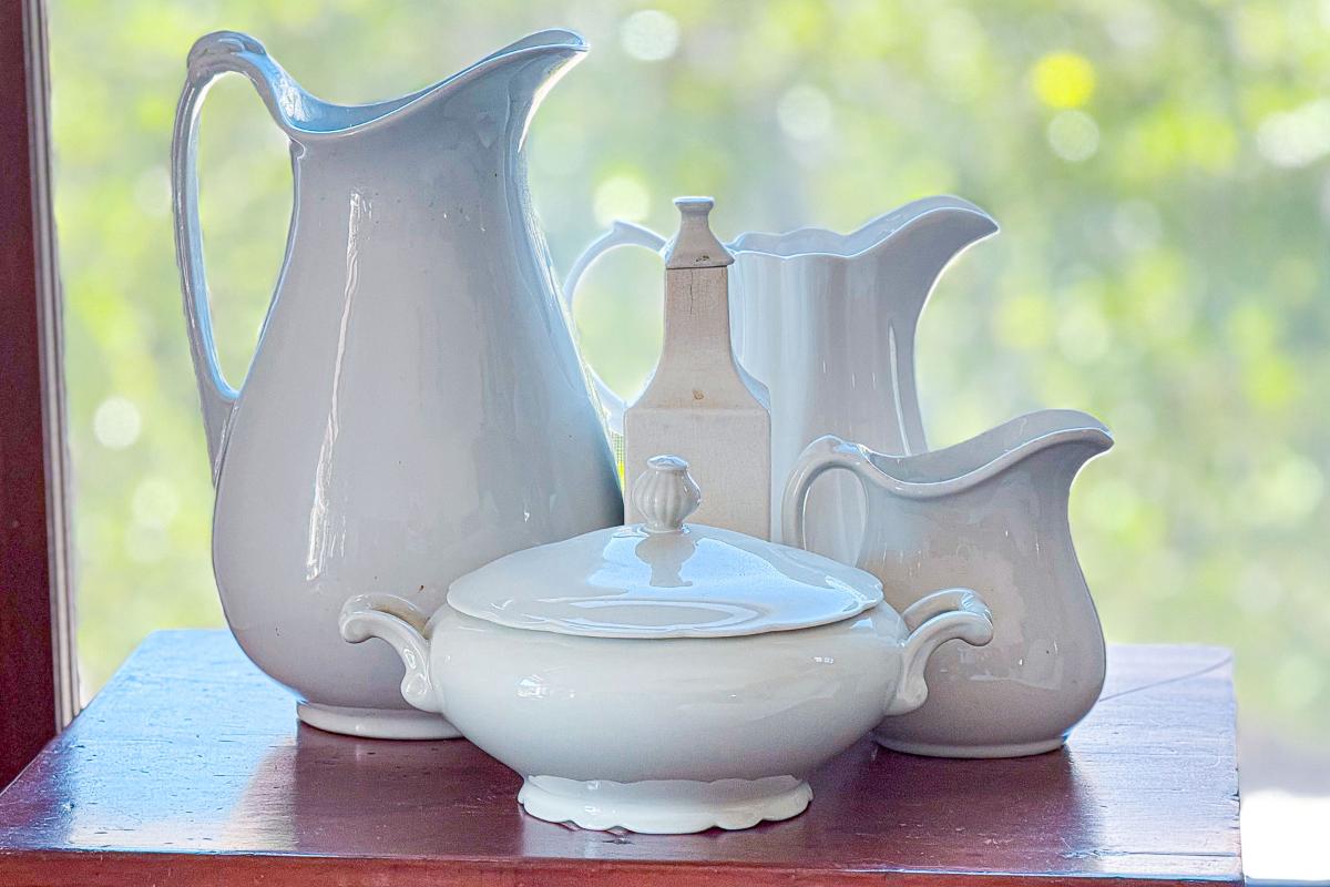 A group of white ceramic pitchers, a lidded tureen, and a tall bottle are arranged on a wooden surface with a blurred green background, suggesting a view through a window. The items vary in size and shape, creating a harmonious display.