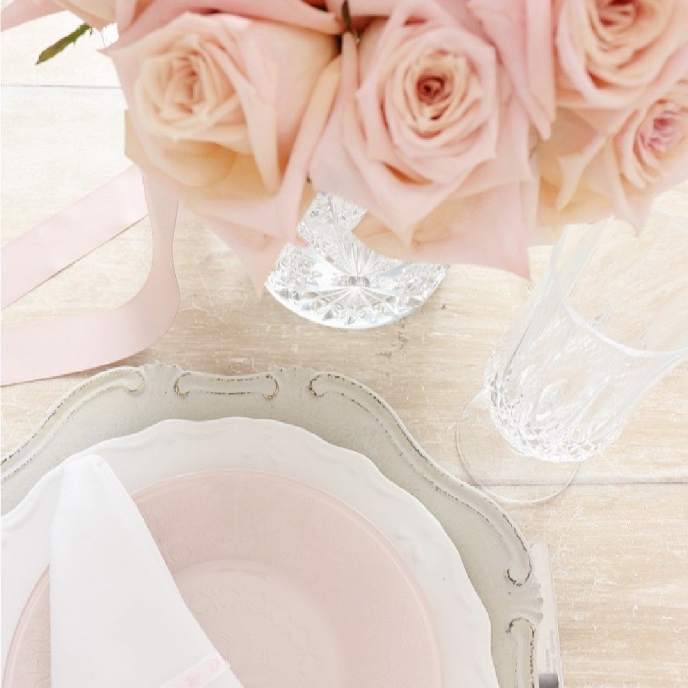 A delicate table setting featuring pink roses in a glass vase, a pink plate with a white napkin atop a larger white plate, and a tall crystal glass on a light wooden surface. Everything is arranged elegantly with soft, pastel colors.