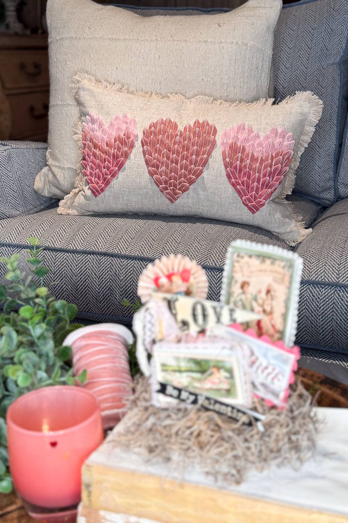 A cozy setting featuring a gray cushion with a pillow adorned with three pink heart patterns. In the foreground, a pink candle and decorative items, including a "Love" sign, sit on a table alongside a small plant.