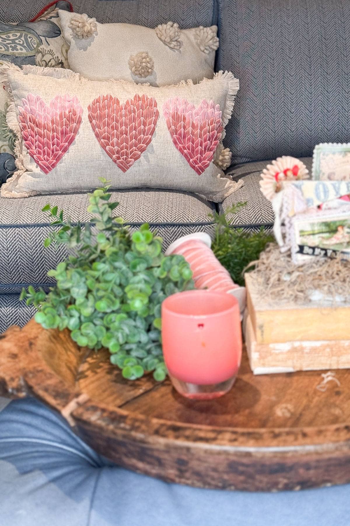 A cozy living room scene with a textured gray sofa featuring a pillow adorned with three pink hearts. A wooden coffee table holds a pink candle, a bundle of greenery, and decorative items in soft, neutral tones.