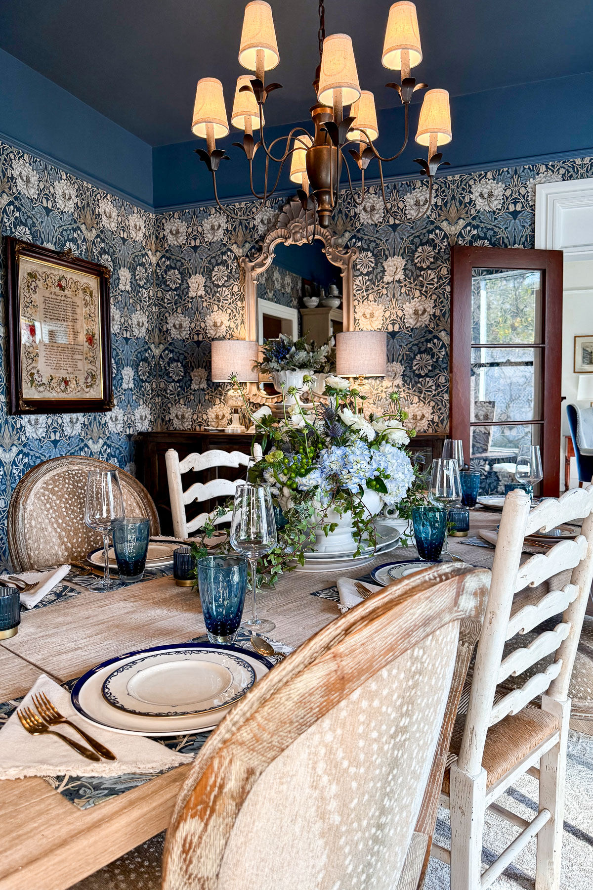 Elegant dining room with blue floral wallpaper, wooden table set for a Sunday Sharing. Blue glassware, white plates, and napkins complement a large floral centerpiece. A chandelier with warm lights hangs above, reflecting softly in the mirror that captures the open door's view.