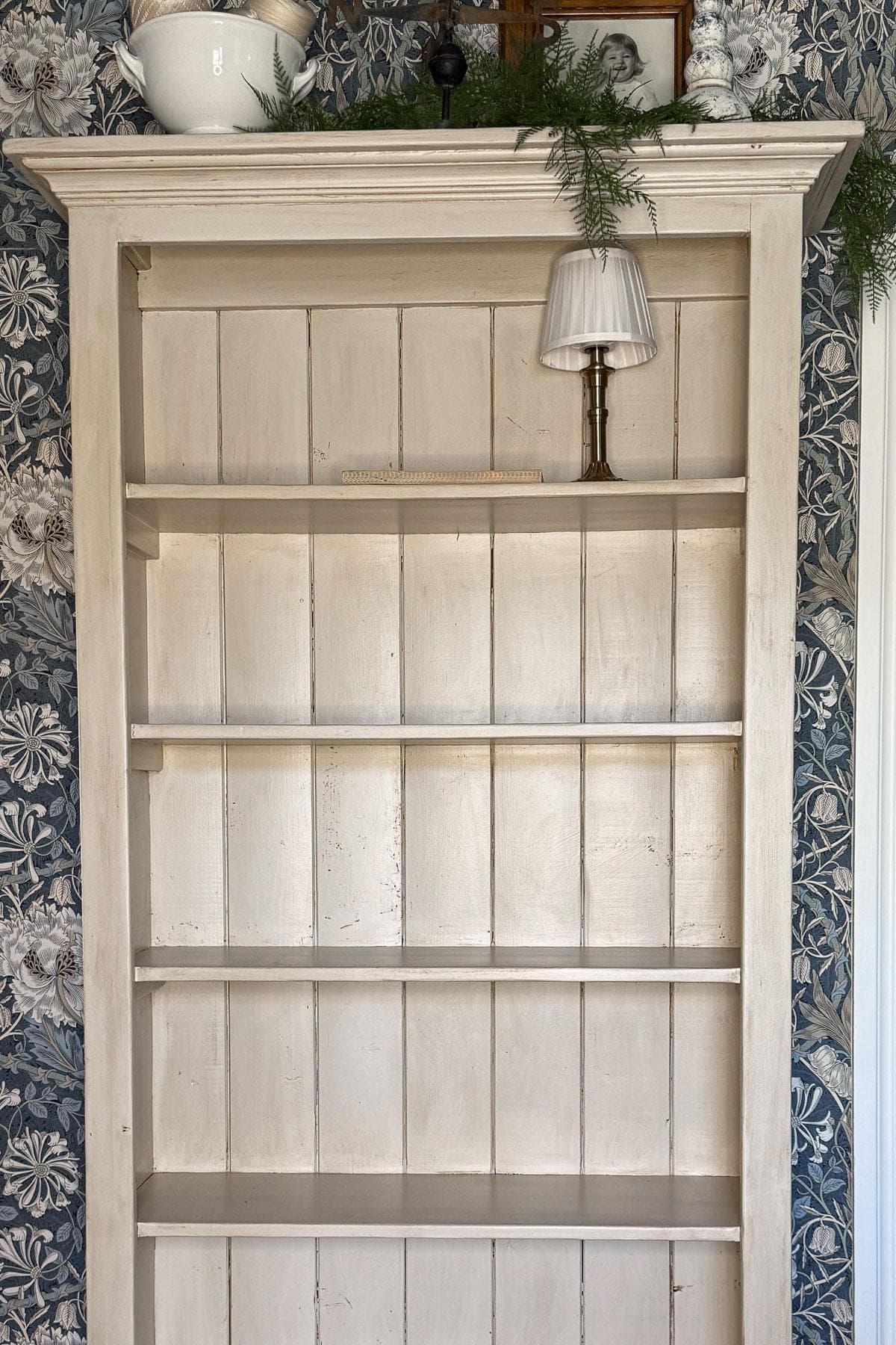 Empty wooden bookshelf with a light cream color stands against a floral-patterned wallpaper in shades of blue and white. A small lamp with a white shade and a bowl with greenery are placed on top of the bookshelf.