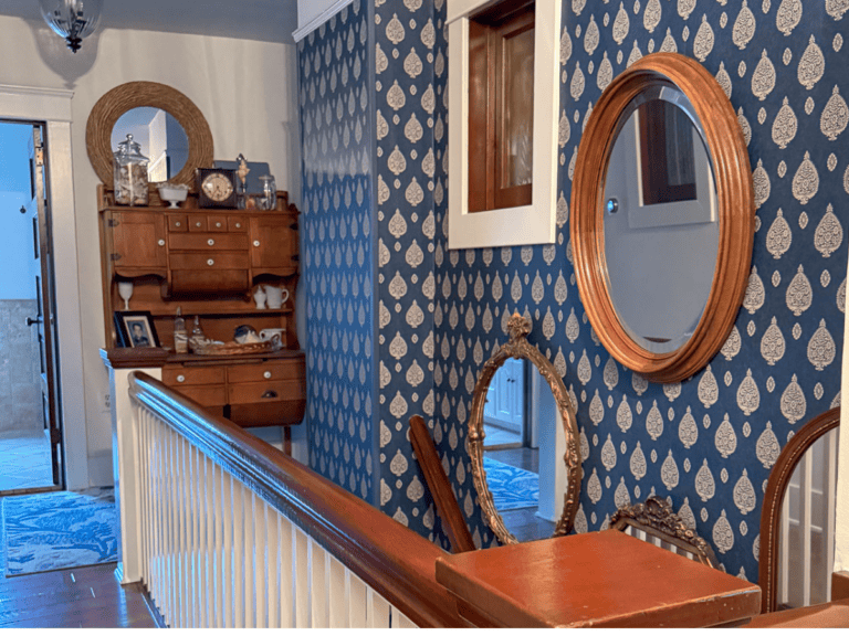 The hallway, resembling a cozy "She Shed," features blue patterned wallpaper and wooden mirrors. A decorative wooden shelf holds various items like a clock and figurines, reminiscent of Sunday Sharing treasures. A wood-trimmed railing graces the staircase, while a rug adds warmth to the floor.