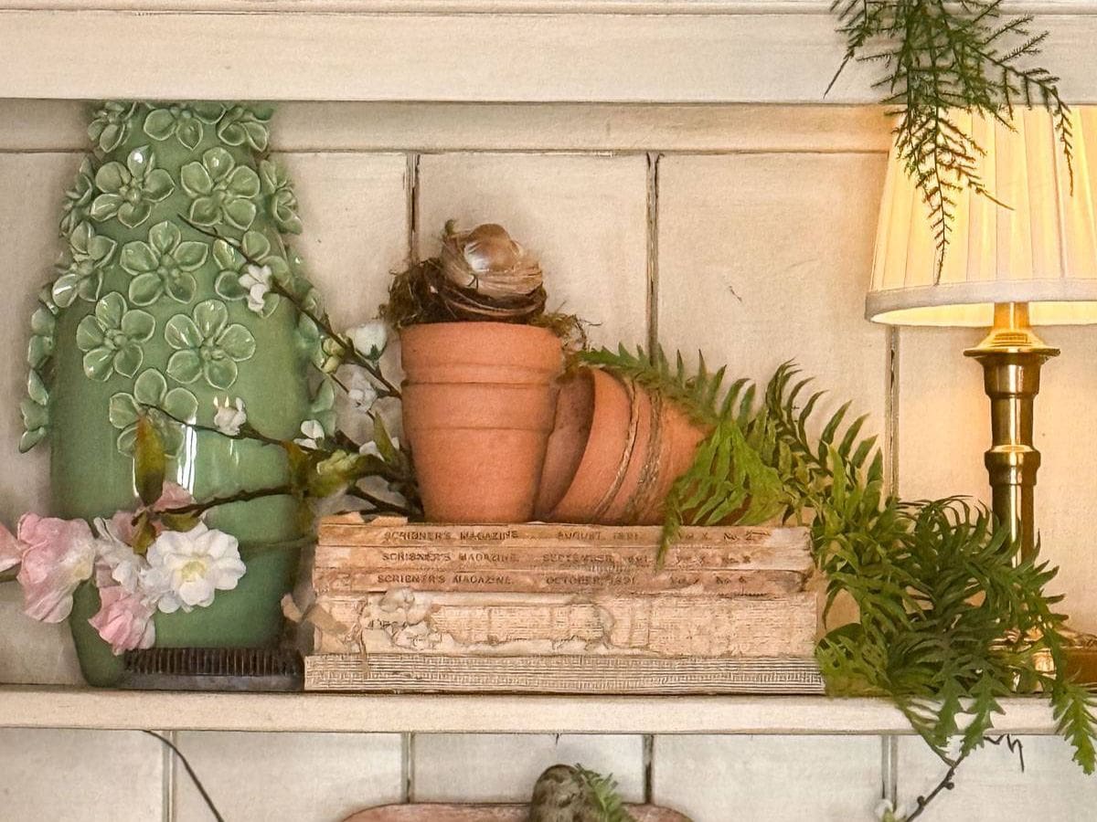 A cozy shelf styled with vintage books, small terracotta pots, and a decorative green tile. A gentle lamp casts a warm glow, while ferns and pink flowers add a touch of nature. Perfect for discovering how to style shelves with timeless elegance.