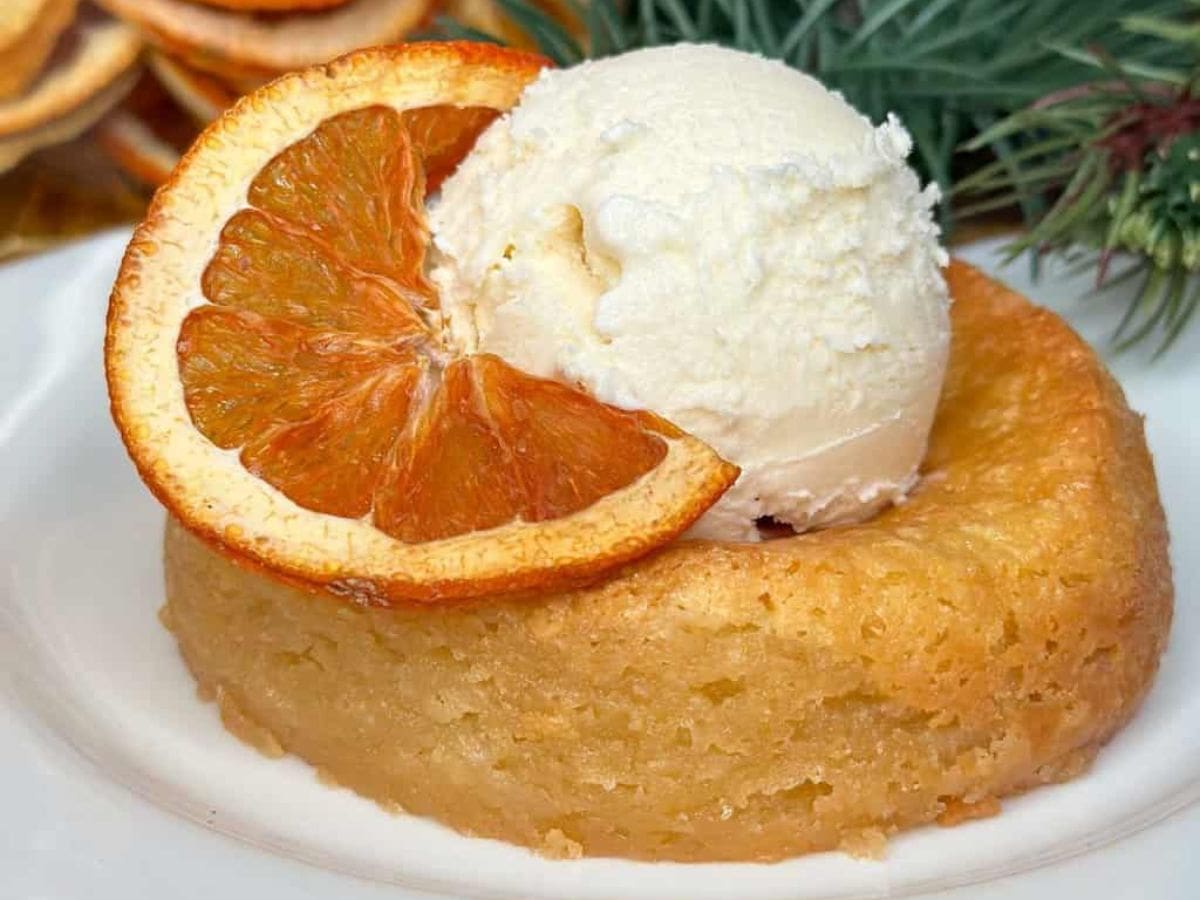 A dessert inspired by Mastro's Butter Cake Recipe features a round cake topped with a scoop of vanilla ice cream, garnished with a slice of dried orange. The cake is light golden brown, surrounded by hints of greenery in the background.