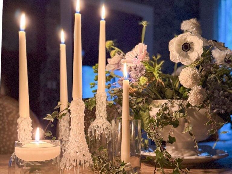 Lit candles of varying heights are elegantly arranged in wine bottle candle holders on a table, with a centerpiece of white flowers and greenery in a white vase. The soft glow creates a cozy atmosphere in the dimly lit room.