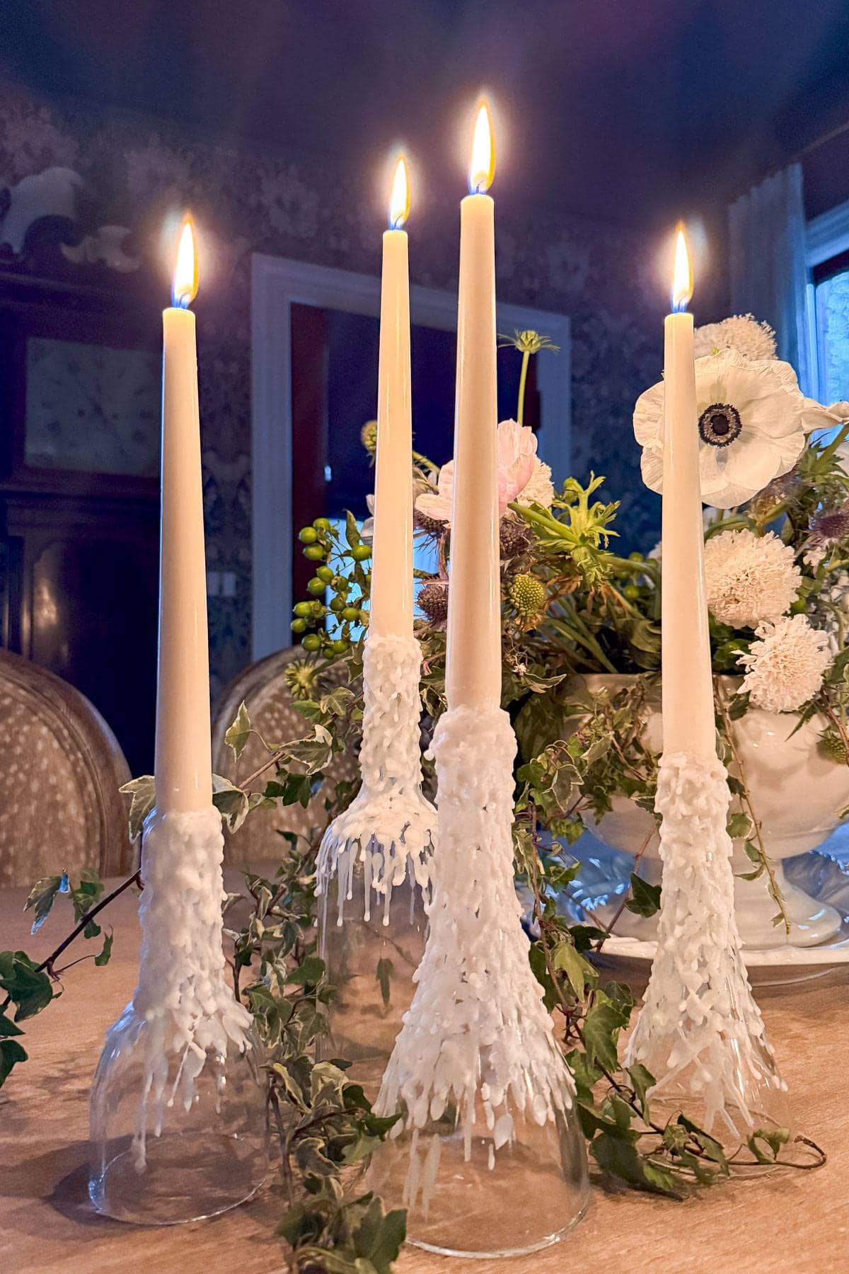 Four lit taper candles in textured holders are arranged on a table, surrounded by greenery and white flowers. The background features a dimly lit room with patterned wallpaper and antique furniture.