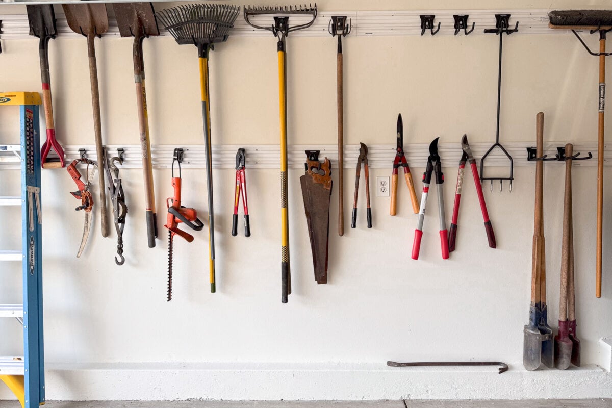 A neatly organized garage wall with various gardening tools hanging on hooks, ready for spring. Tools include rakes, shovels, a saw, clippers, and a leaf blower. A blue ladder is partially visible on the left side as you start preparing for the season.