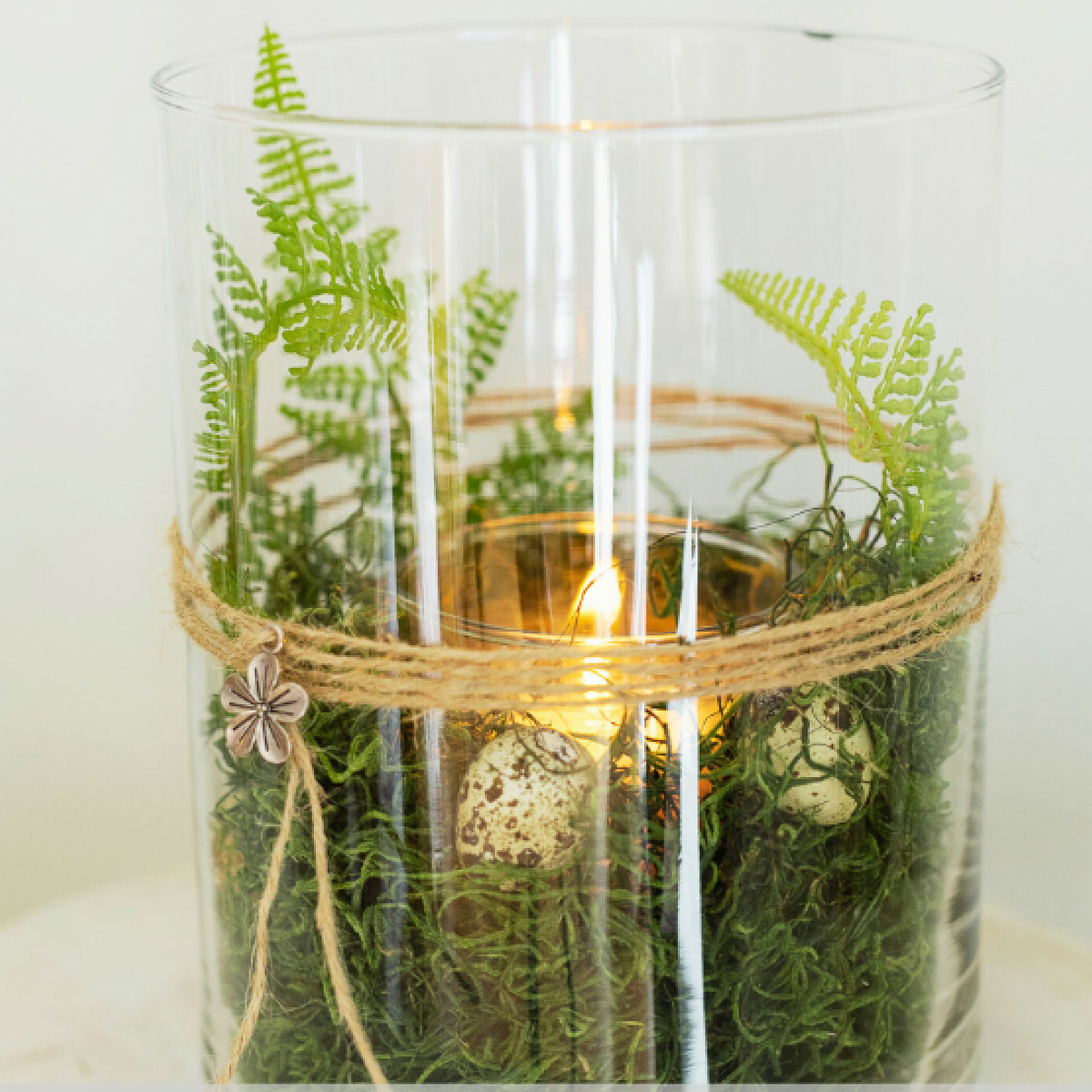 A glass vase contains green moss, ferns, and a small candle. Two decorative eggs rest among the greenery. Twine with a star-shaped charm is wrapped around the vase.