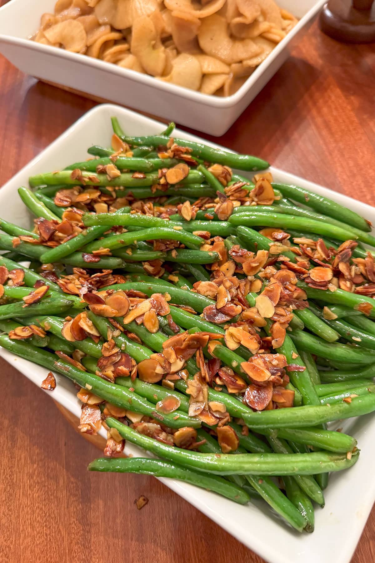 A rectangular white dish filled with sautéed green beans topped with toasted almond slices. In the background, there's another dish containing cooked pasta shells. Both dishes are on a wooden table.