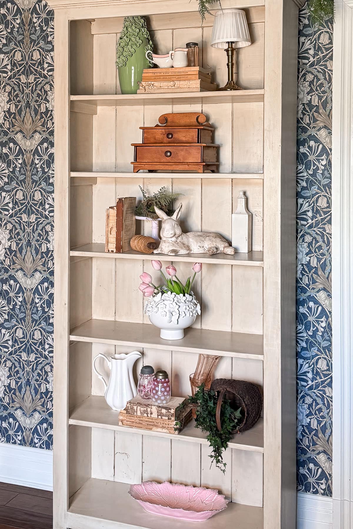 Learn how to style shelves with this beige wooden bookshelf. It boasts decorative items like books, a bunny sculpture, a lamp, vases of pink tulips, candy jars, rustic boxes, and a white ceramic pitcher—all set against a blue floral-patterned wallpaper for added charm.