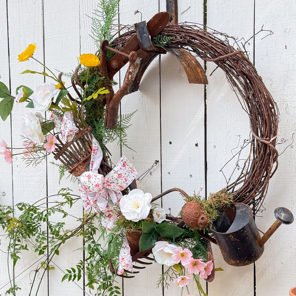 This Hand Crafted Society decorative wreath is made of intertwined branches on a white wooden background. It features pink and yellow flowers, small gardening tools, a floral bow, and a miniature watering can as accents.