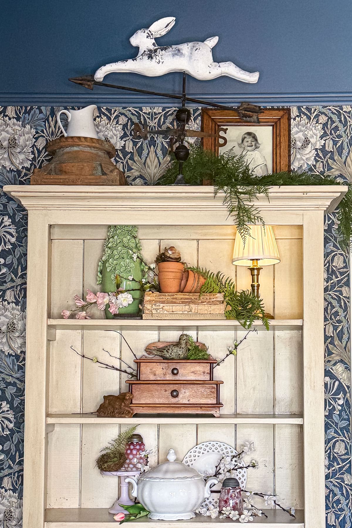 A decorative shelf, styled to perfection, features a vintage rabbit figurine, framed photo, potted plants, a small lamp, and charming vintage items. The floral wallpaper offers a classic touch while greenery drapes gracefully across the shelf.