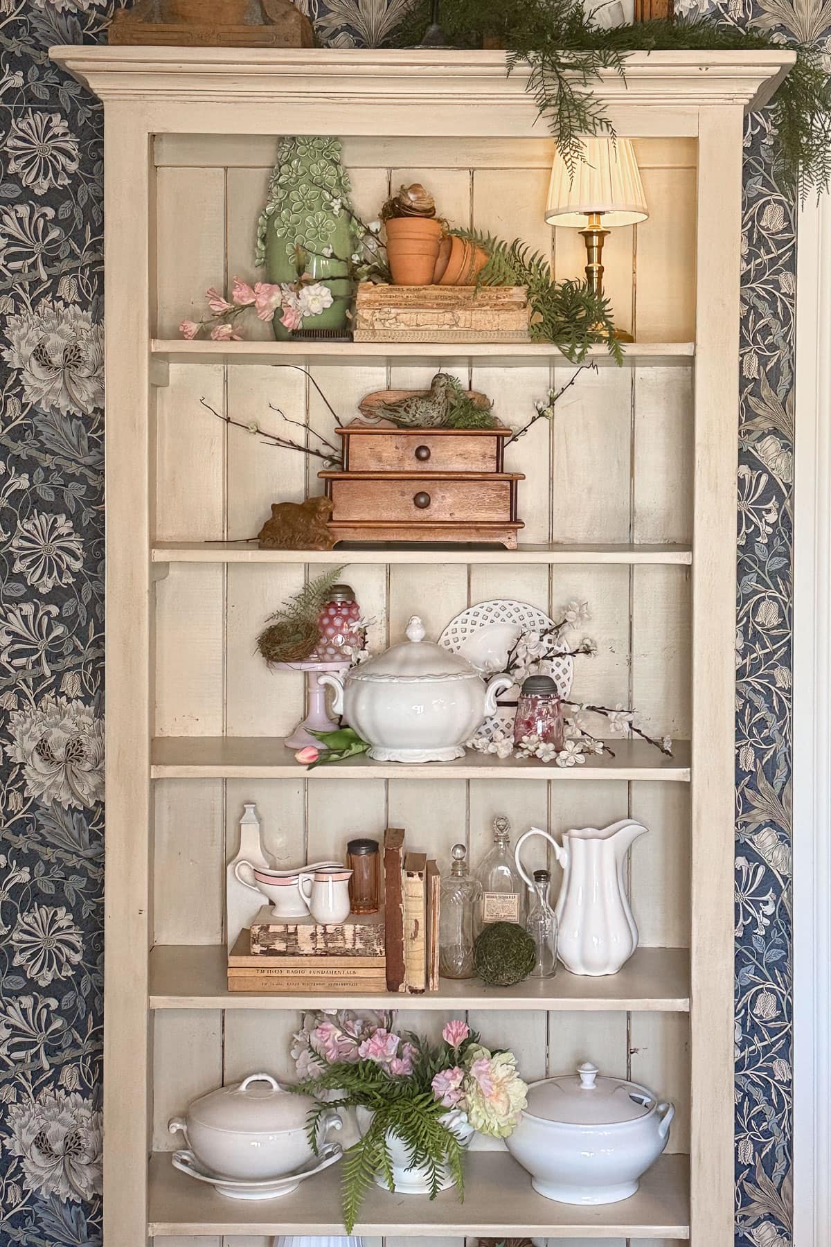 Discover how to style shelves with a cream-colored wooden design, adorned with white teapots, a wooden jewelry box, and assorted trinkets. Enhance the charm with a small lamp and potted plants against floral wallpaper.