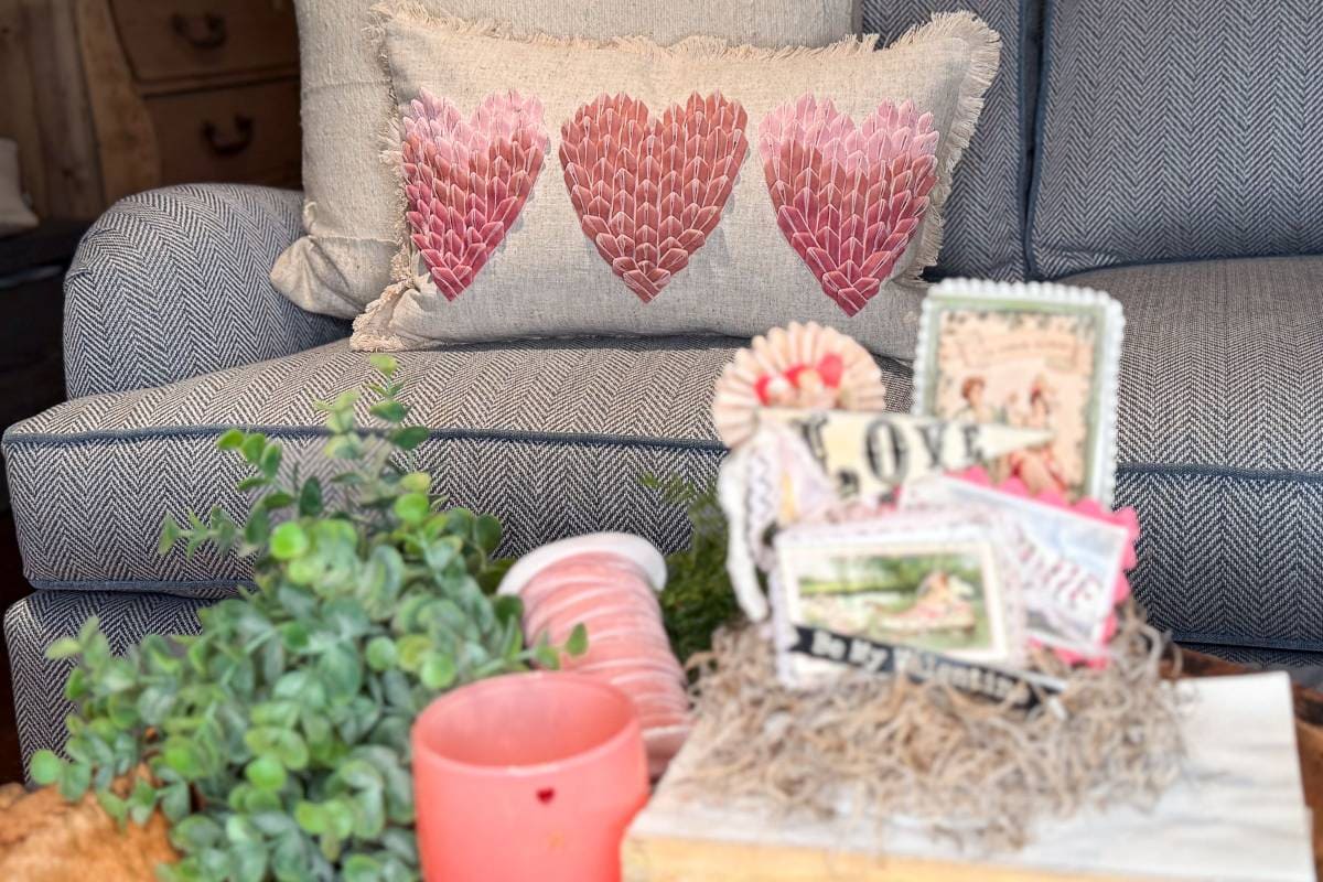 A cozy setting with a blue herringbone-patterned sofa featuring a decorative pillow adorned with three pink heart designs. In the foreground, there's a table with a potted plant, a red candle, and a small basket filled with ribbon and vintage-themed items.