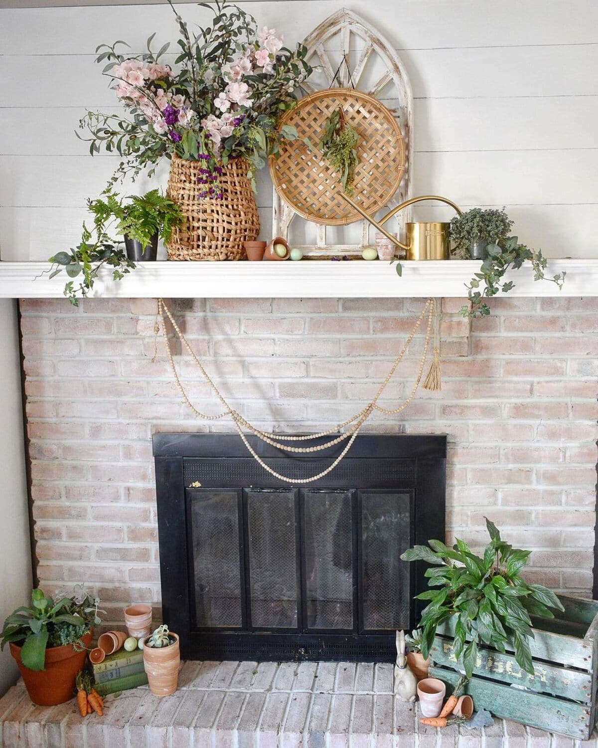 A white brick fireplace with a black metal screen is adorned with a variety of potted plants and decorative items. A wicker basket, a golden watering can, and a latticework piece sit on the mantel. Strings of beads hang elegantly across the front.