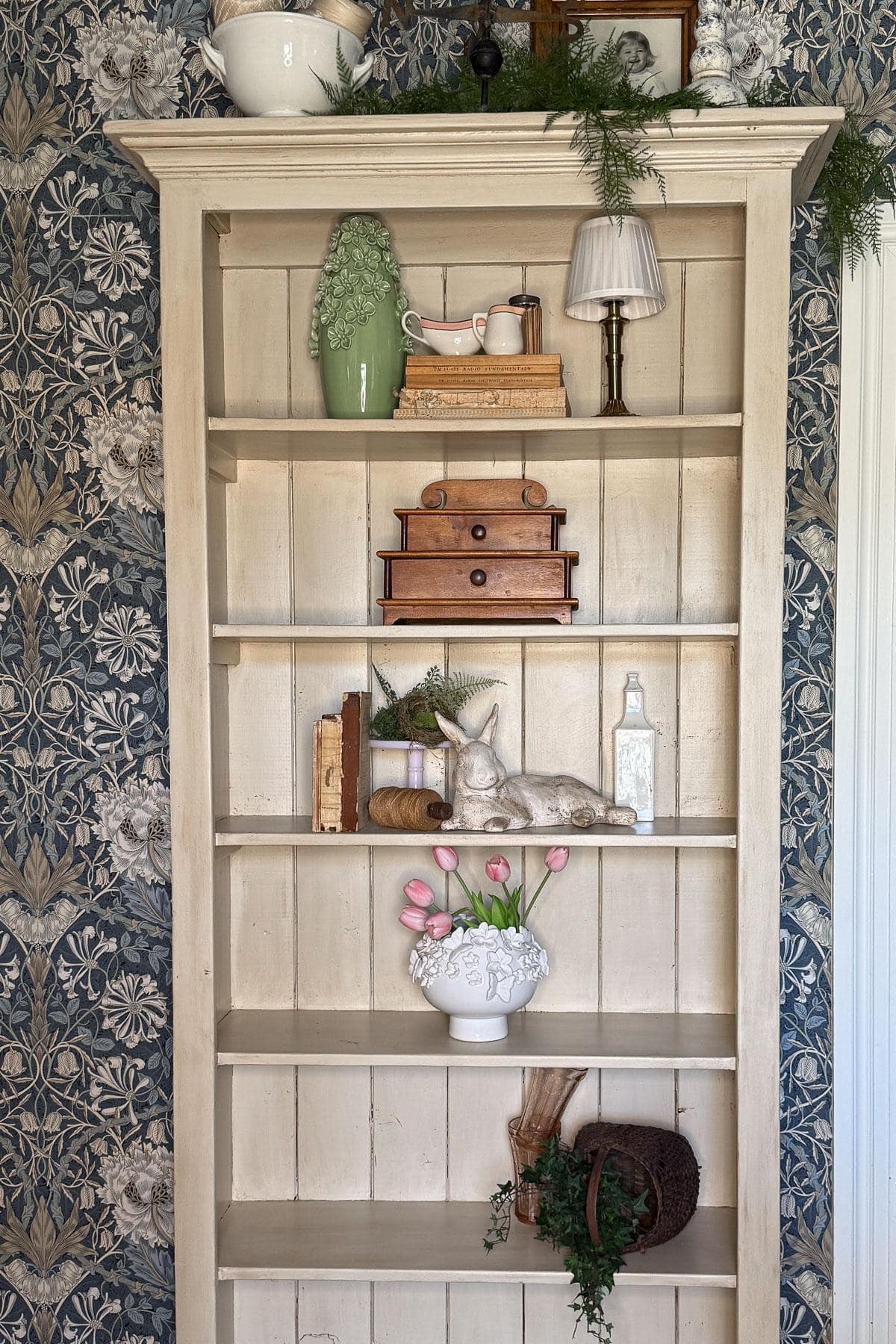 A cream-colored wooden shelf against floral wallpaper. The shelves hold a mix of vintage items: a green ceramic pitcher, stacked books, a wooden box, a rabbit figurine, a vase with pink tulips, and some greenery.