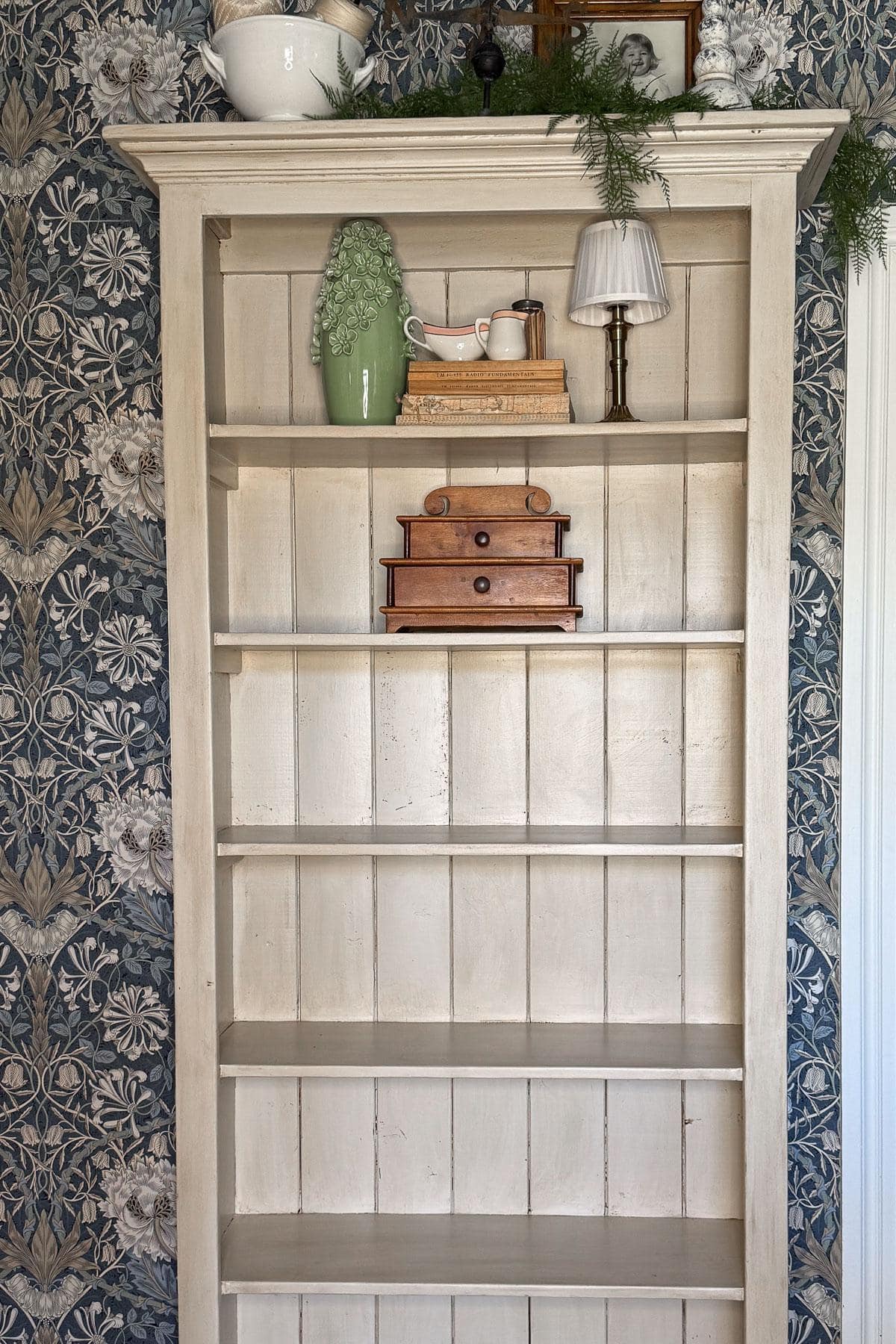 A rustic wooden shelf partially filled with decor items including a small green Christmas tree, books, a vintage tea set, a wooden jewelry box, and a lamp. The shelf is set against floral wallpaper. Greenery and a photo frame adorn the top.