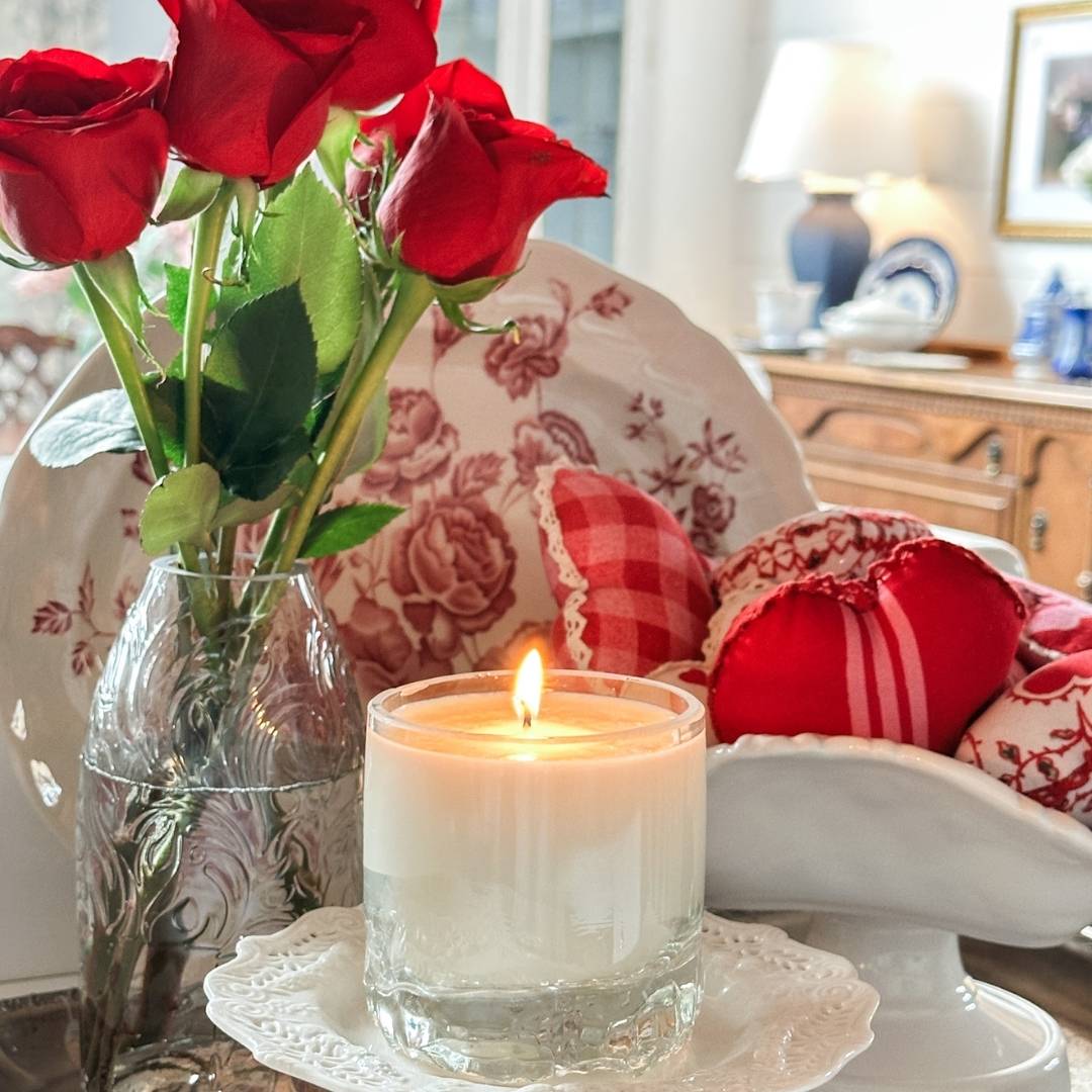 A glass candle burns on a saucer beside a vase of red roses and a decorative bowl filled with heart-shaped fabric ornaments. The background shows a wooden cabinet with a lamp and framed picture.