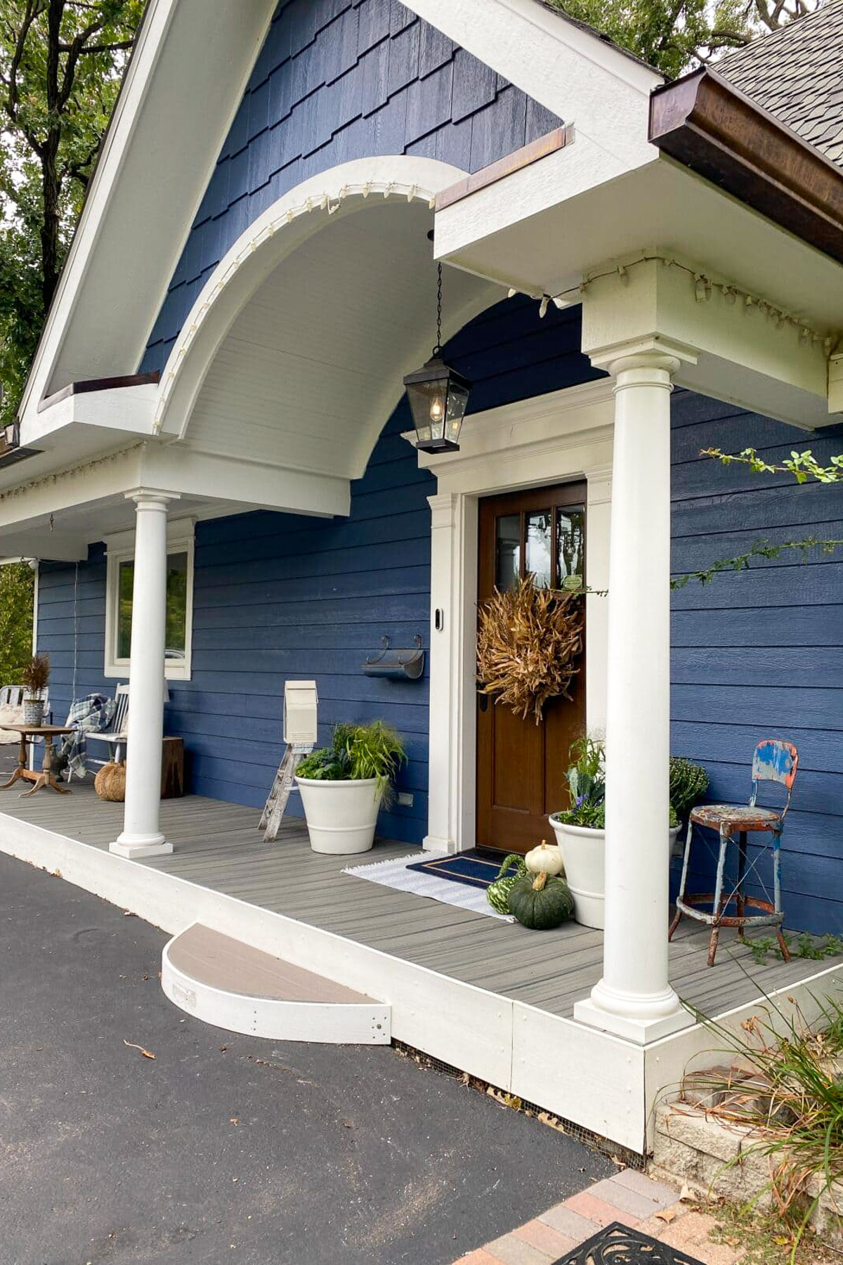 A charming house exterior with blue siding and a white, arched porch invites you to explore. White columns support the roof, while a wooden door is adorned with a wreath. Potted plants and a vintage chair decorate the porch, providing an adventure in elegance as you step toward the driveway.