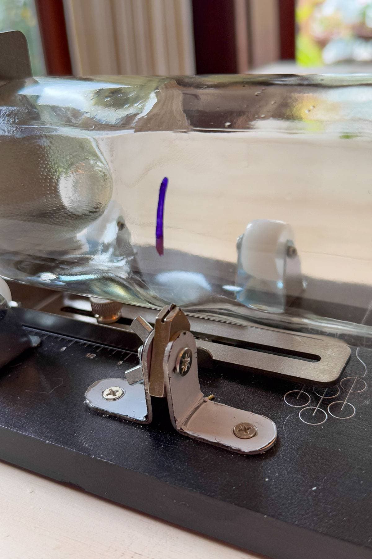 Close-up of a glass tube apparatus with a metal component on a black base. The tube contains a small, glowing purple line. Natural light filters from the background, highlighting the reflective surfaces.