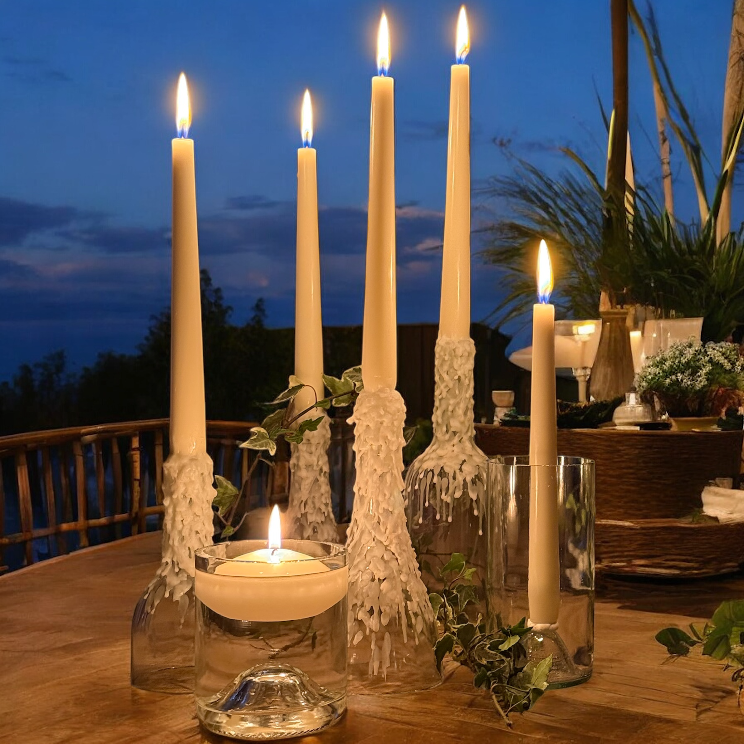 Tall candles with wax drippings stand on a wooden table, surrounded by greenery and framed by DIY wine bottle candle holders. A smaller candle flickers in front. The background reveals a darkening sky with clouds, hinting at nightfall, with a glimpse of outdoor greenery and furniture.