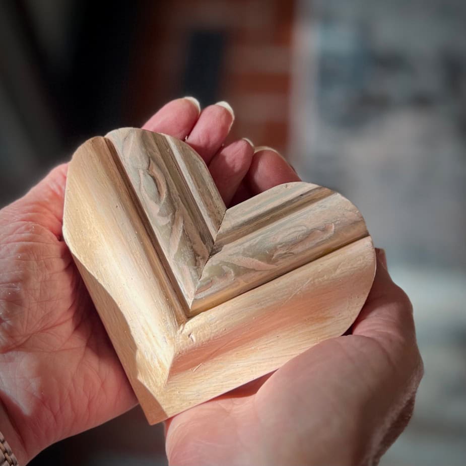 Hands holding a carved wooden heart with a textured surface, intricate patterns adorning its surface. The background is softly blurred, drawing attention to the heart—a perfect piece for Sunday Sharing in your cozy She Shed sanctuary.