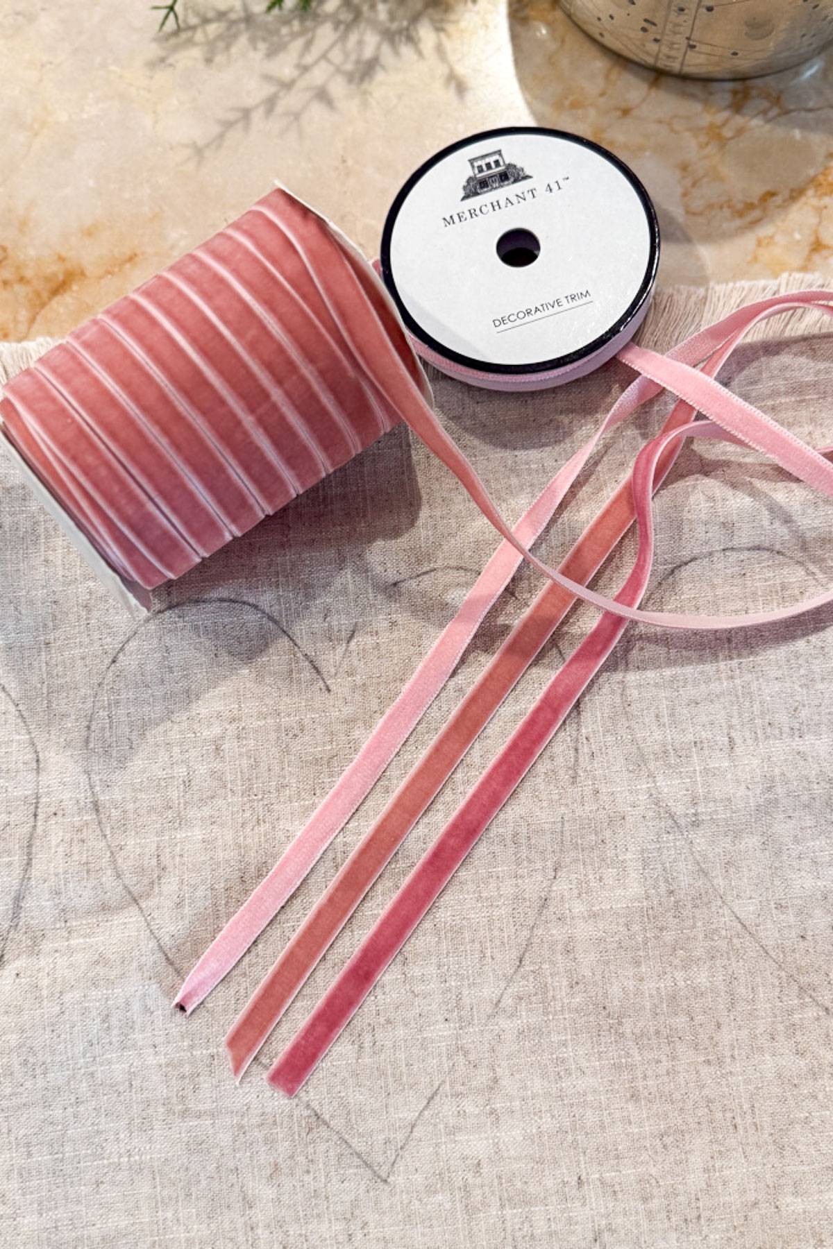 A spool of pink decorative ribbon lies on a piece of fabric. Several strands of the ribbon are pulled out and arranged neatly. The fabric has a faintly drawn heart shape on it, and a potted plant is partially visible in the background.