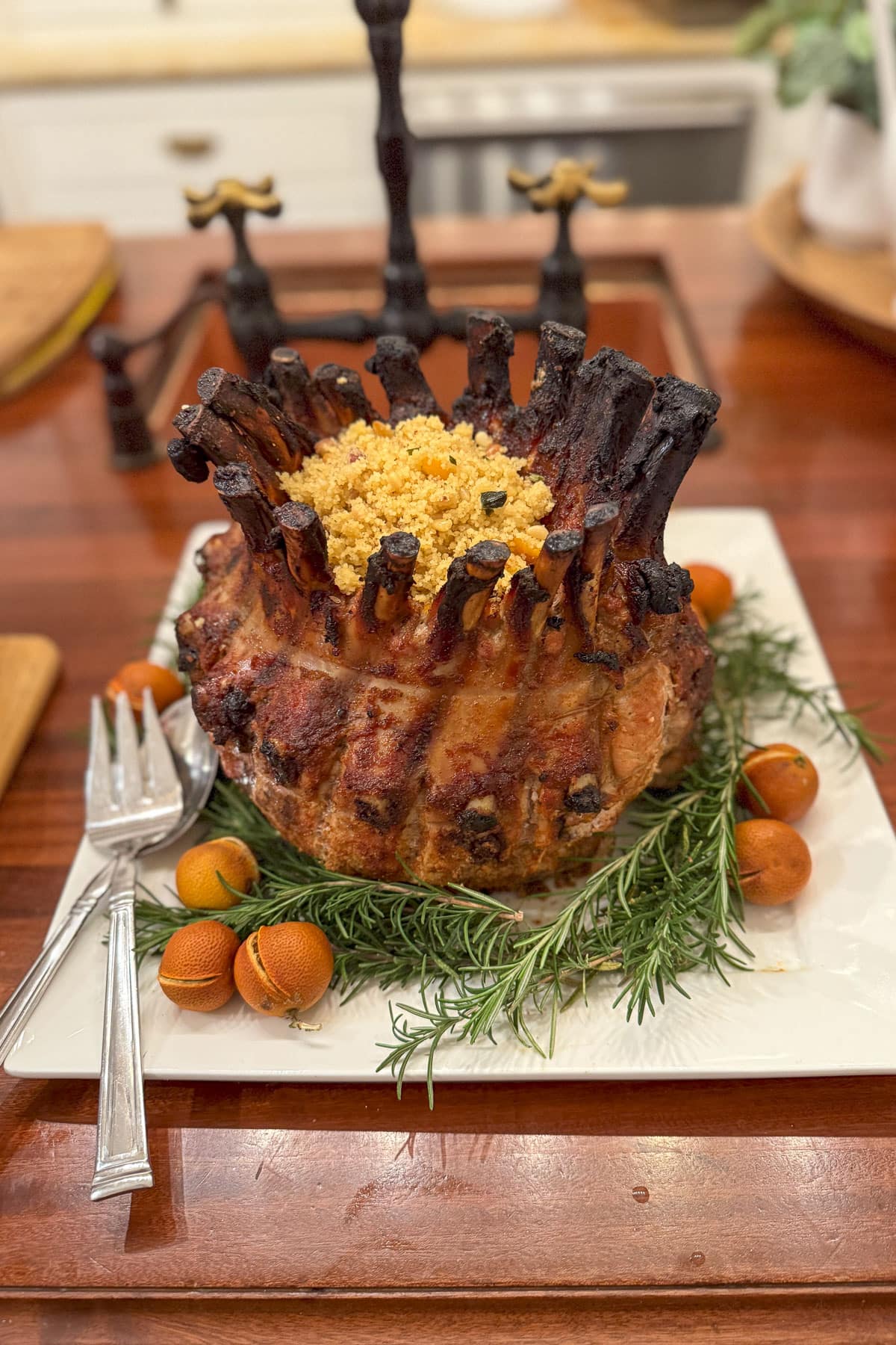 A cooked crown roast with crispy bones on a white platter, garnished with fresh rosemary and small oranges. The roast is stuffed with a breadcrumb mixture, and two silver serving utensils are placed alongside it.