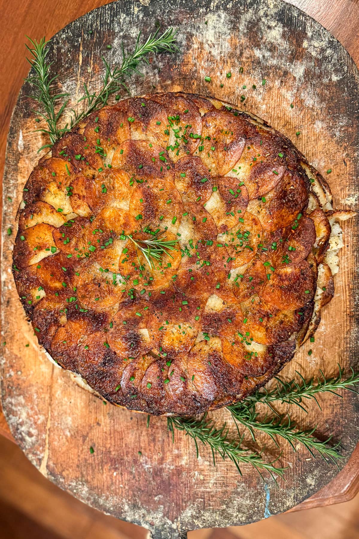 A golden-brown potato galette garnished with fresh rosemary and finely chopped chives, presented on a rustic wooden board.