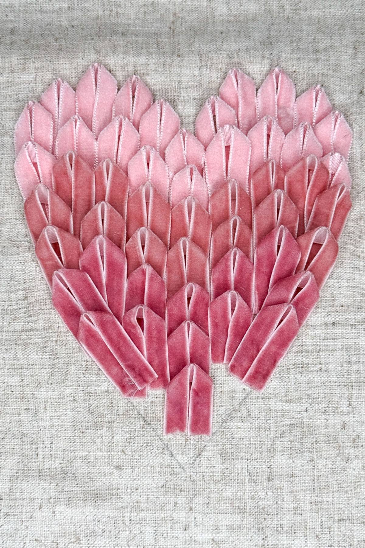 A heart-shaped design of pink and red fabric folds arranged in gradient shades, creating a textured pattern on a light beige background.