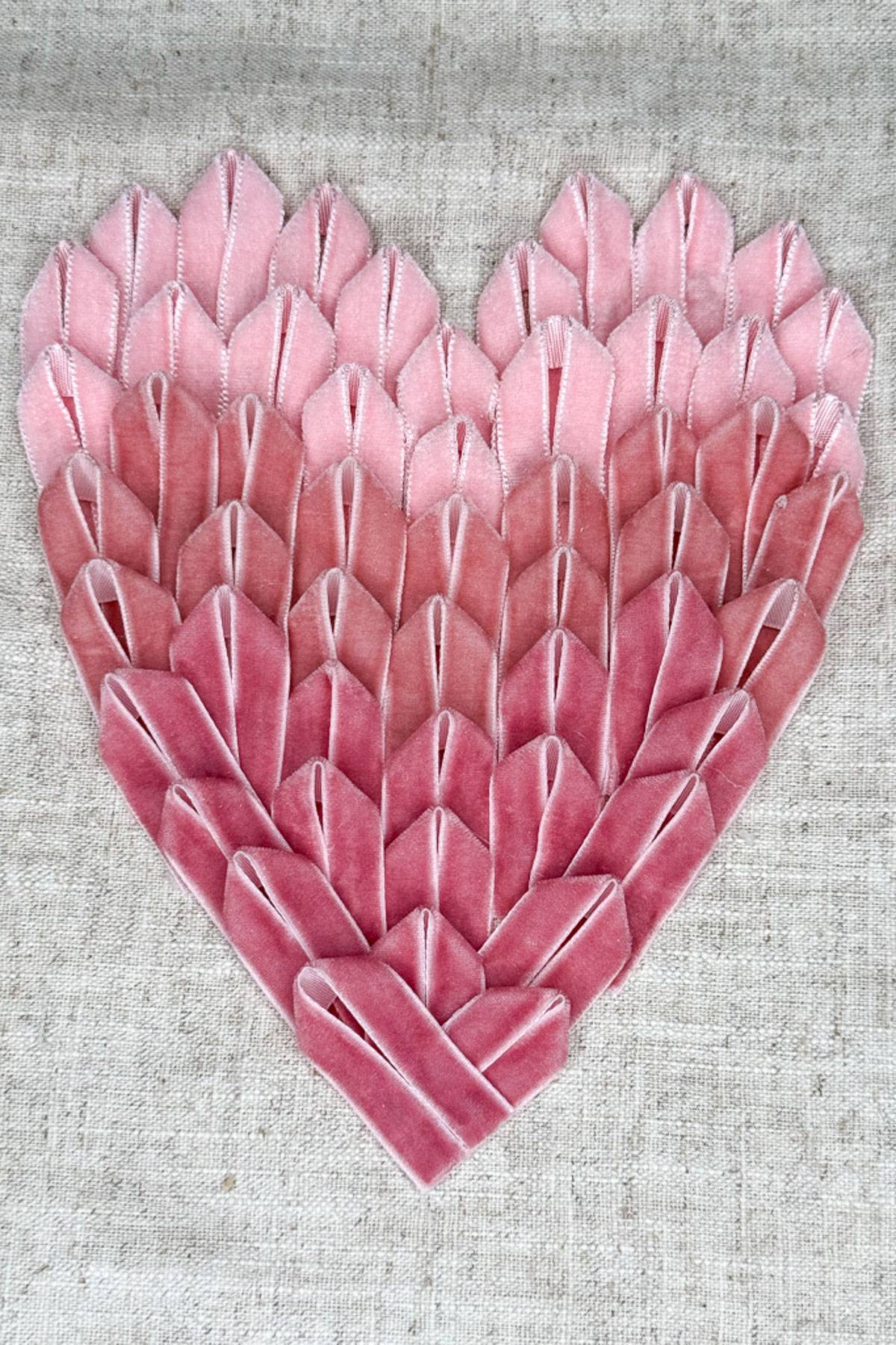 A heart shape crafted from folded pink ribbons is displayed on a textured beige background. The ribbons create a layered, textured pattern, with colors transitioning from light to dark pink from top to bottom.