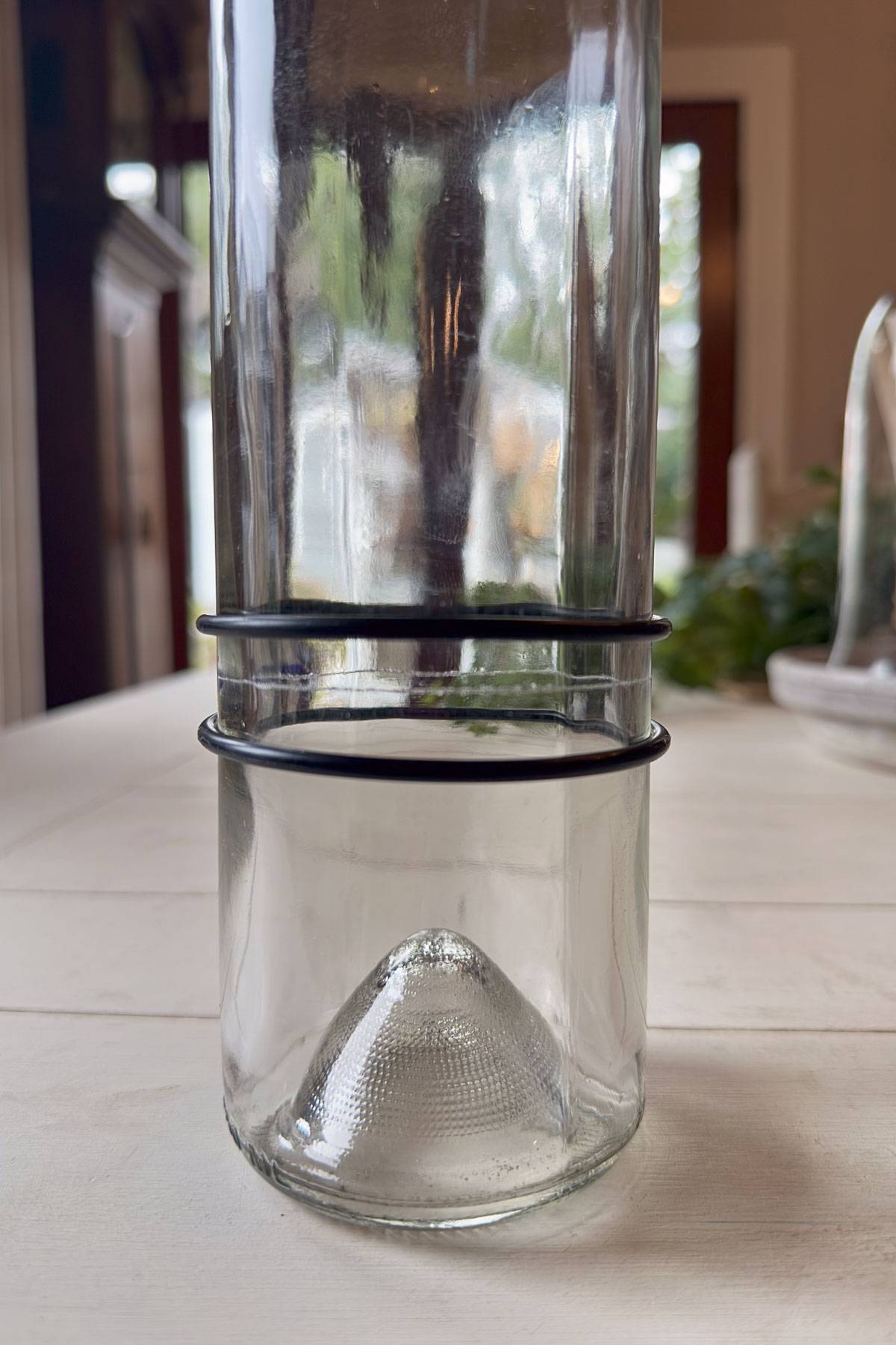 A clear glass bottle with a narrow neck and a cylindrical base rests upright on a wooden table. Two black rubber bands are around the neck and the base. A blurred background shows a window and greenery.