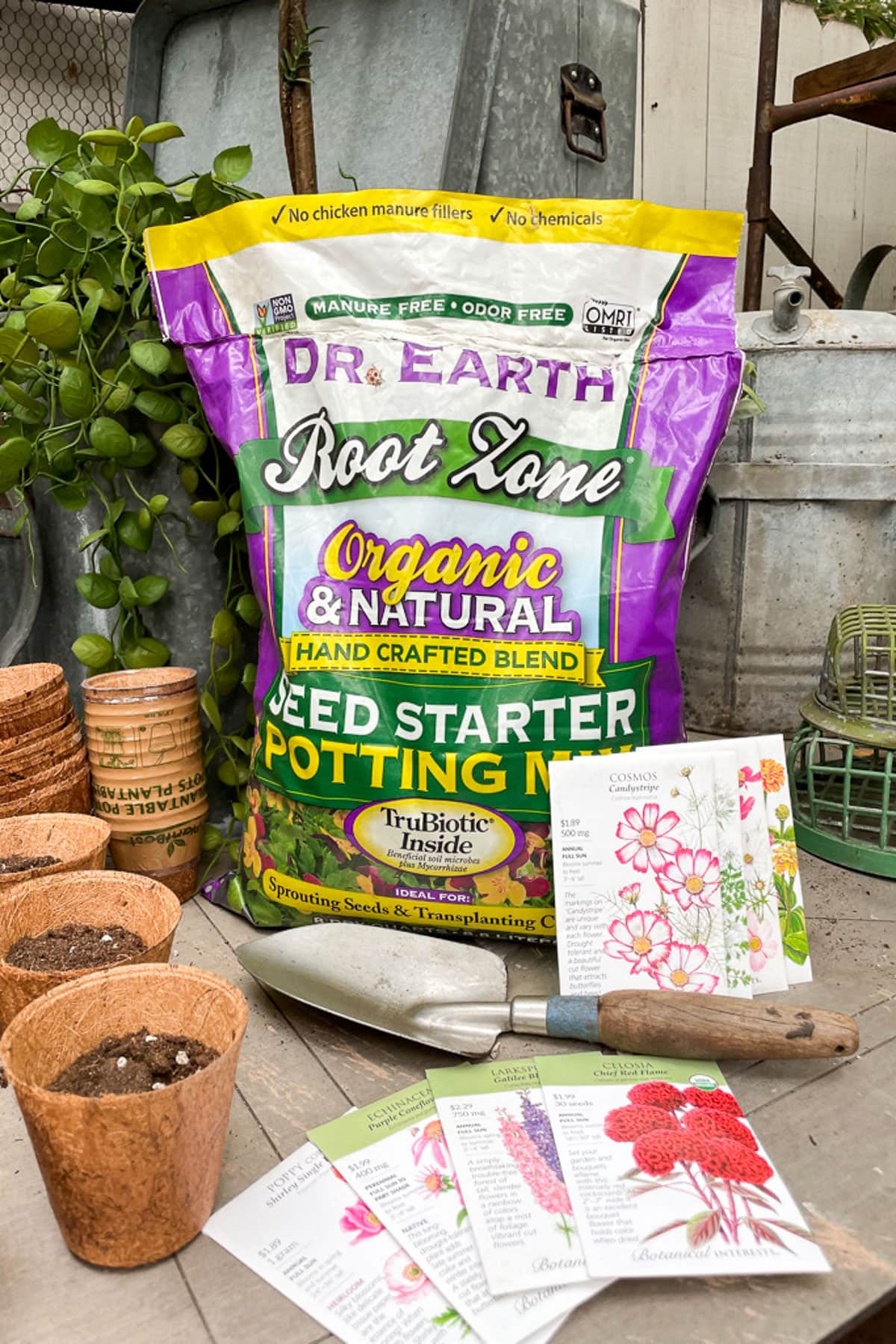 Preparing your garden for spring-A bag of Dr. Earth Root Zone organic seed starter mix is displayed with peat pots, soil, and various seed packets featuring flowers. A small shovel is also visible, set in a garden setting.