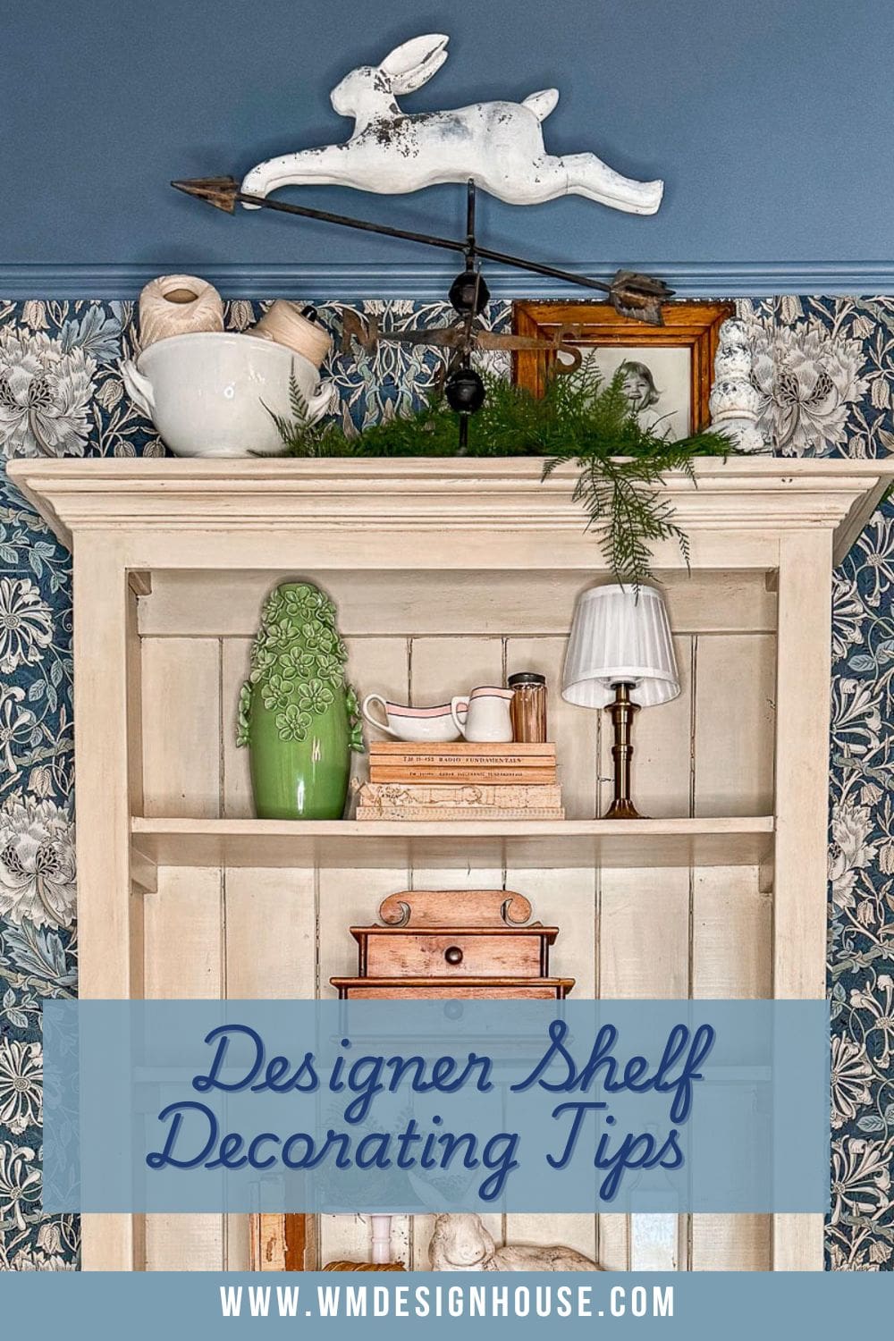 A white wooden shelf with a decorative rabbit on a stick, vases, books, a lamp, and greenery. The wall behind has floral wallpaper and a blue paint. Text at the bottom reads: "Designer Shelf Decorating Tips" with a website URL.