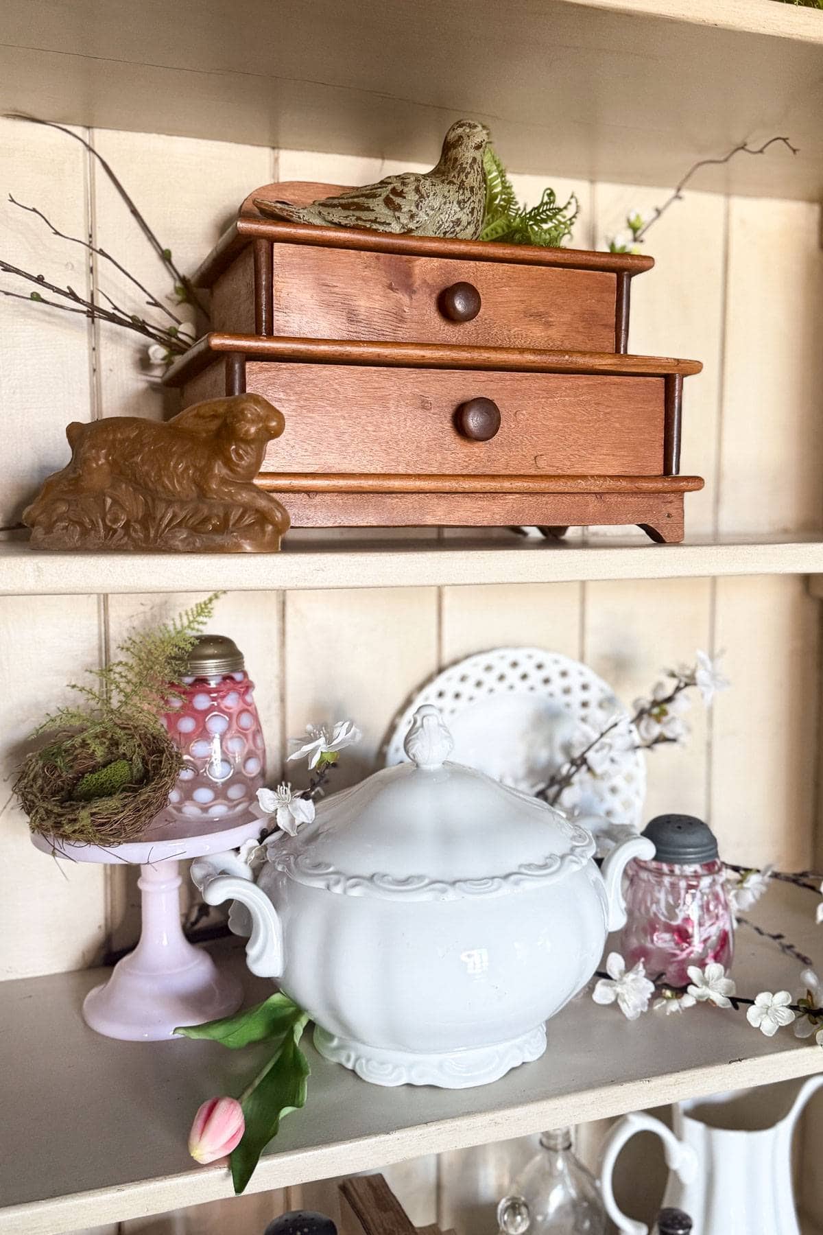 A decorative shelf with a small wooden chest, ceramic bird, and lamb figure on top. Below, a white ornate bowl with lid, glass jars, a pink vase with a bird's nest, and a pink tulip. Delicate branches and a polka-dot plate adorn the background.