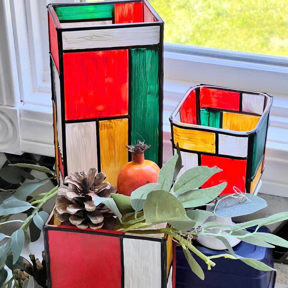 Two stained glass vases with red, yellow, green, and white panels sit on a windowsill. A pine cone, pomegranate, and eucalyptus leaves are nearby, with sunlight streaming through the window in the background.