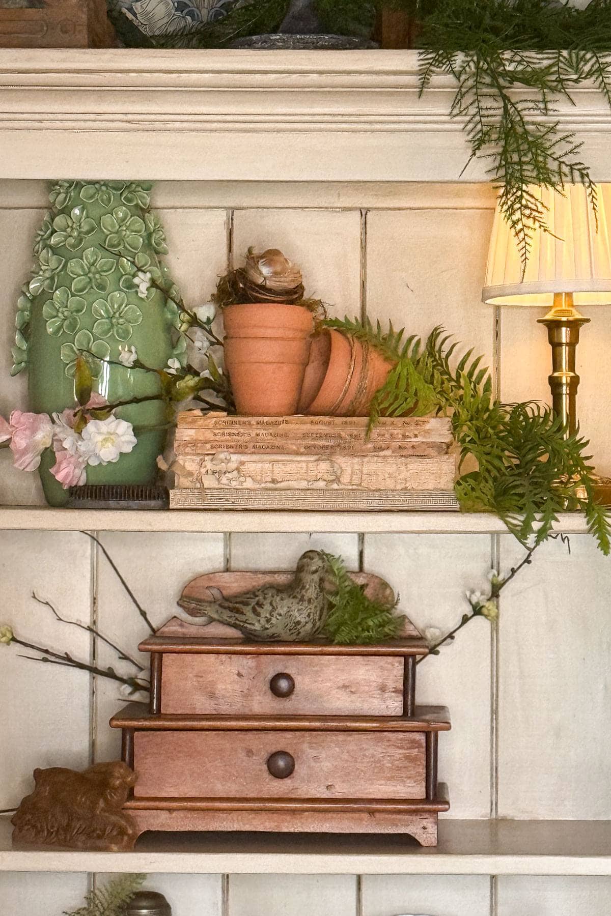 A rustic shelf with stacked books, terracotta pots, green ferns, and a small lamp. A decorative piece featuring flowers and a bird rests on a miniature wooden chest with drawers. The setting is cozy and nature-inspired.