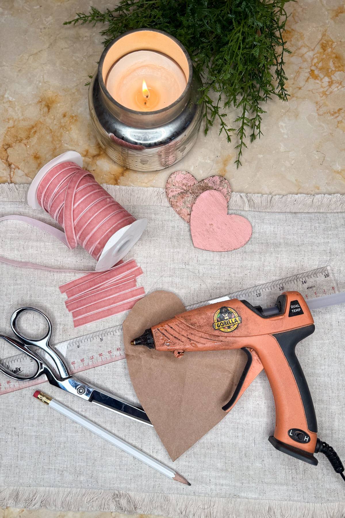 Crafting supplies on a table include a pink ribbon spool, scissors, a glue gun, ruler, pencil, and cutout hearts. A lit candle and small green plant sit in the background, all arranged on a light fabric cloth.