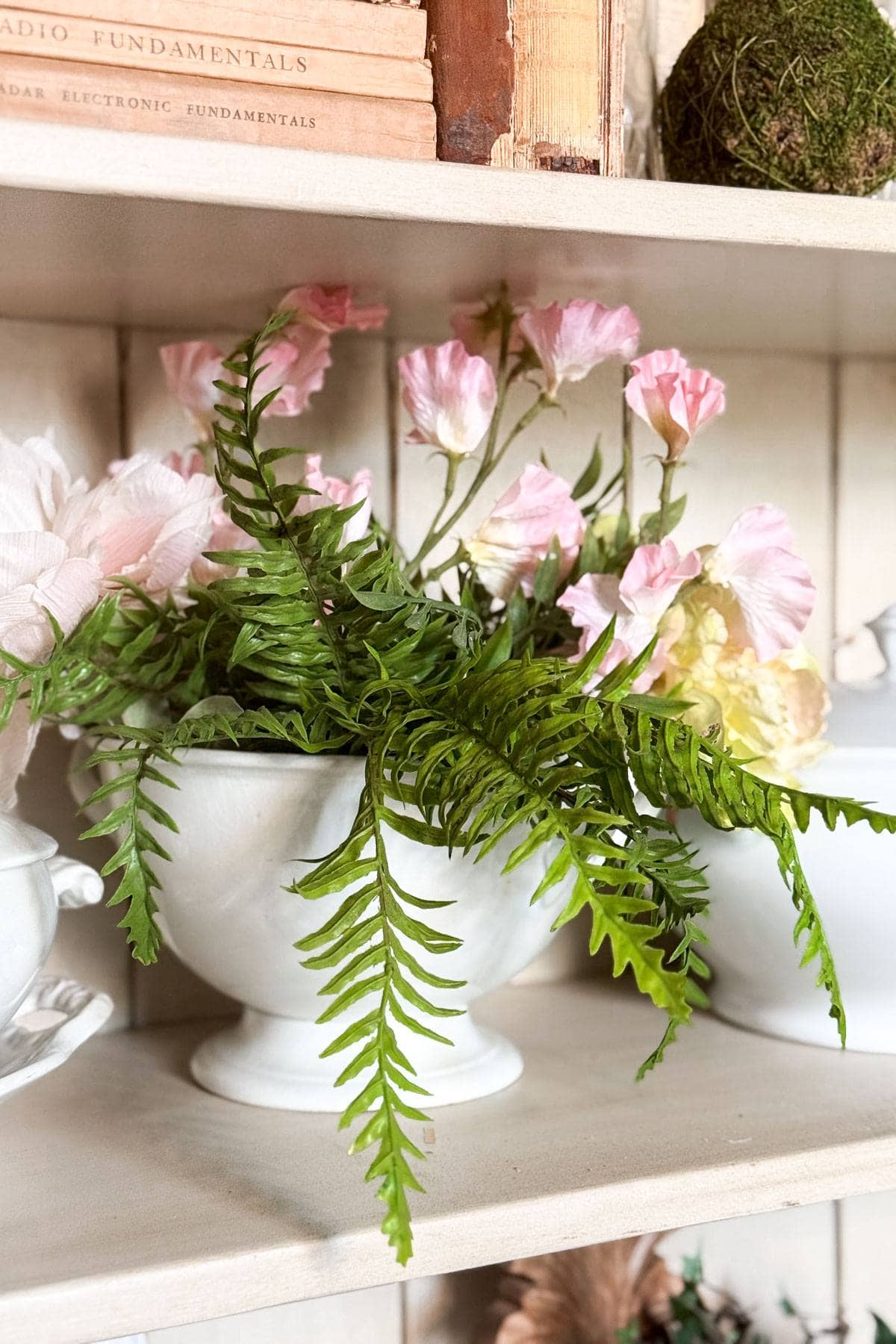 A white ceramic bowl on a shelf holds pink flowers and lush green fern fronds. Books with faded spines are stacked above, and a small moss ball decorates the shelf. The background is a light wood panel.